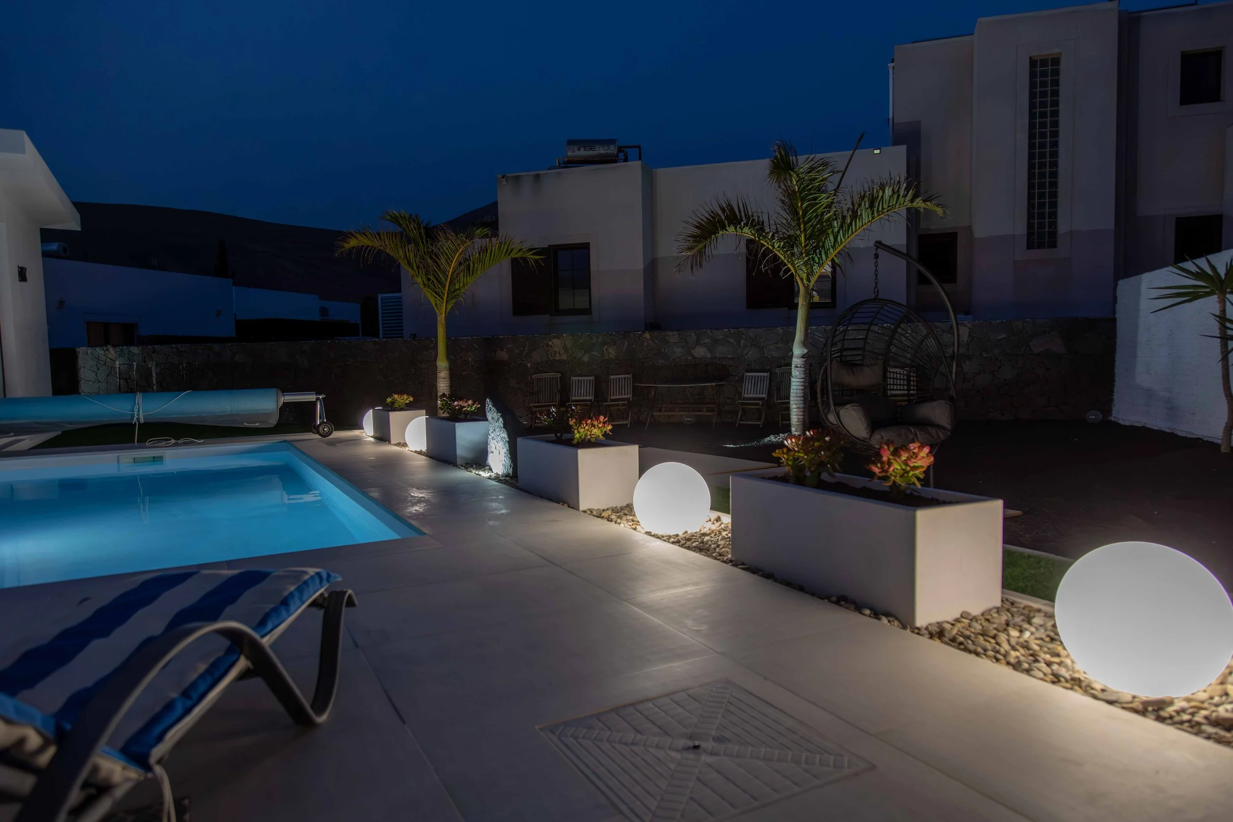 Night view of a modern backyard with a swimming pool, illuminated globe lights, two palm trees, and outdoor lounge chairs.