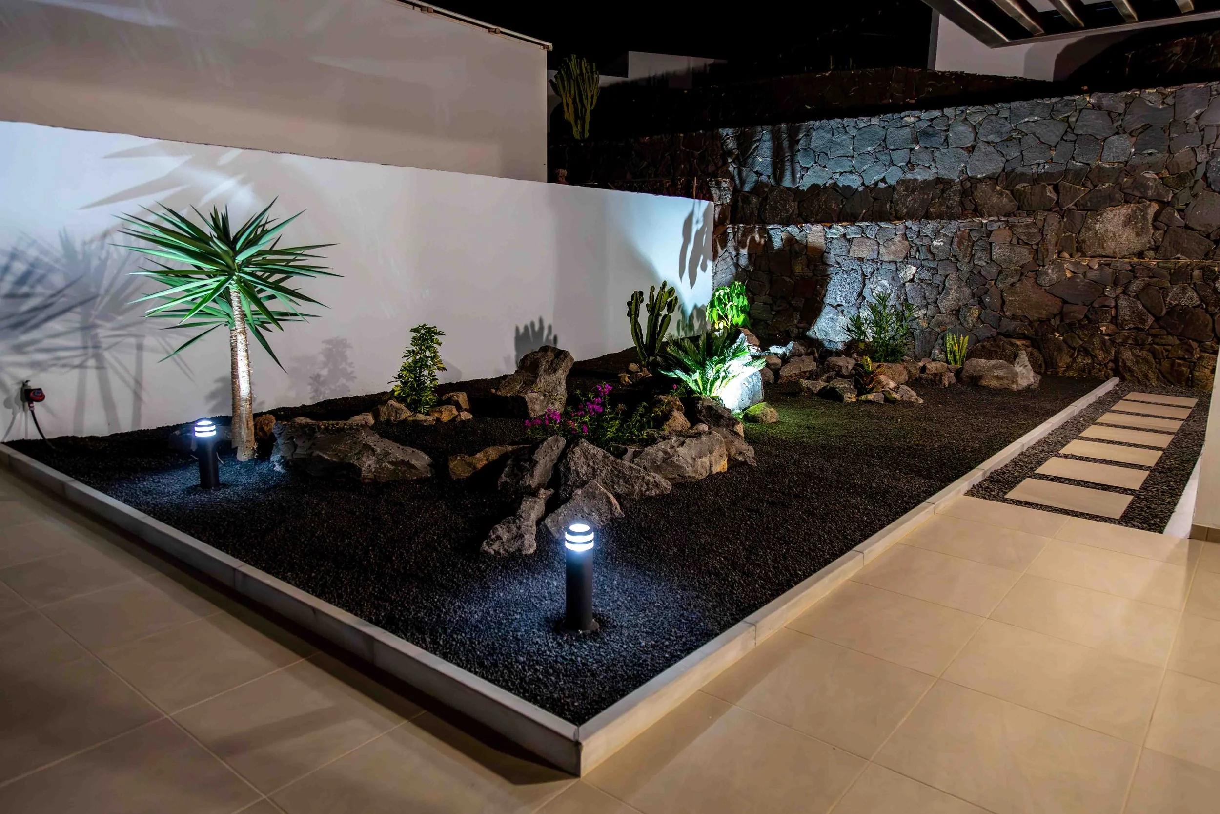 Nighttime view of a modern garden with various plants, rocks, and pathway lights, enclosed by a white wall and a rock wall.