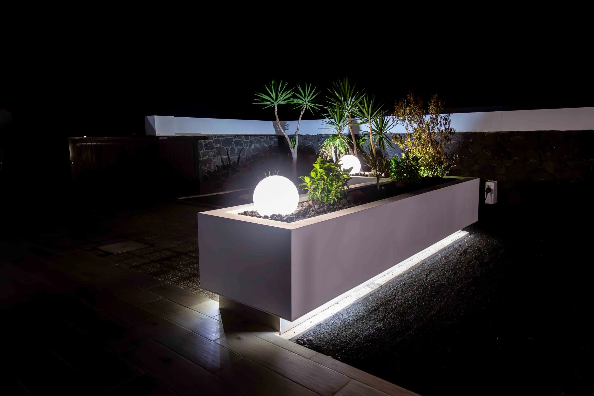 Night view of a modern outdoor garden with illuminated spherical lights, green tropical plants, and a stone wall.