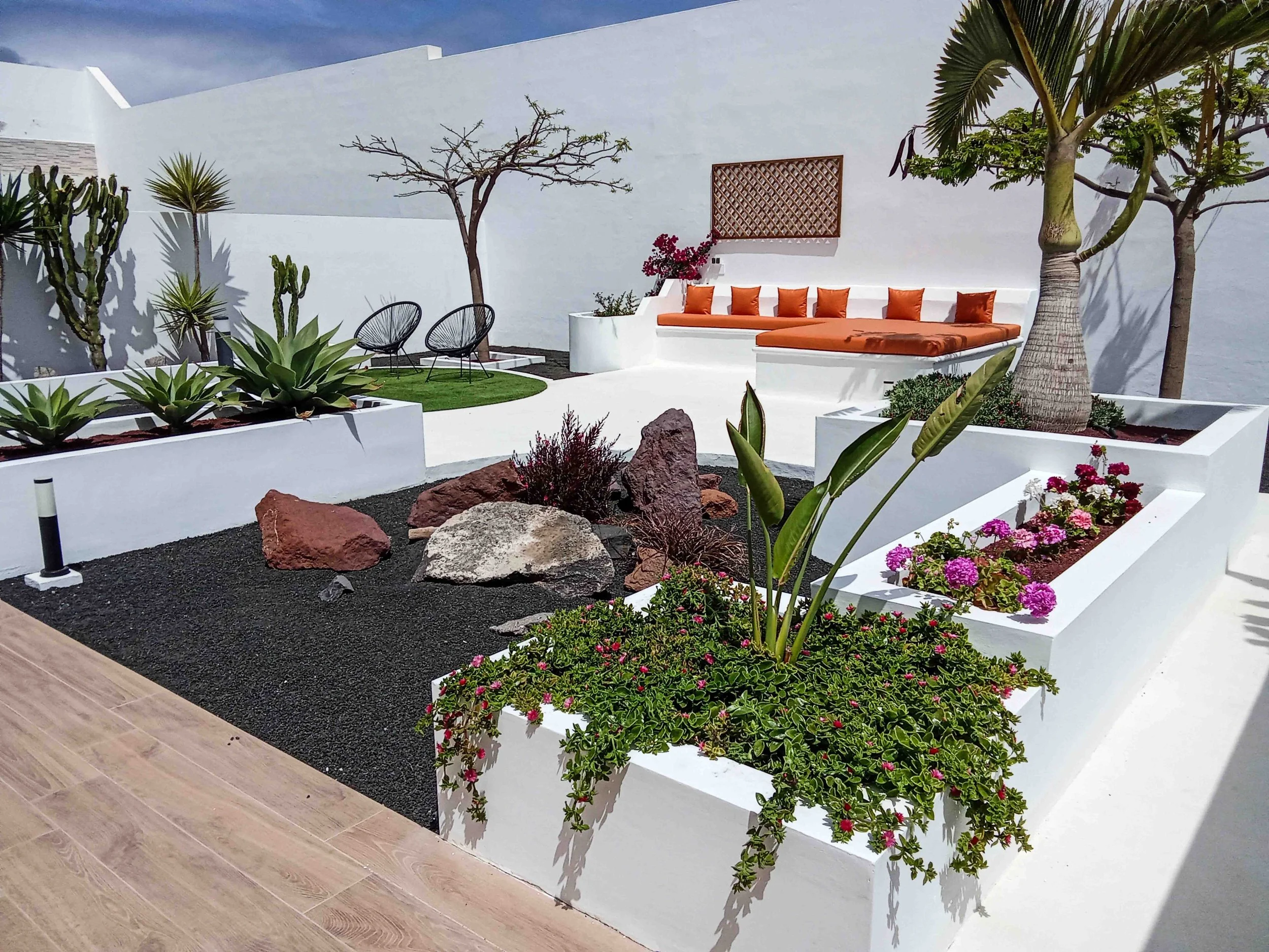 A modern outdoor patio with white walls and planters, orange seating, and a variety of green and flowering plants, including cacti, succulents, and palm trees, with a clear sky in the background.
