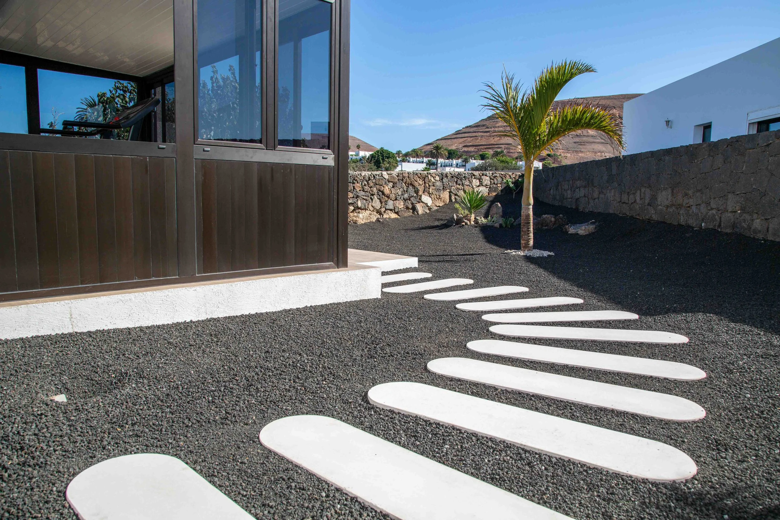 Pathway with white stepping stones leading to a white building with a brown wooden patio, a small palm tree, and a stone wall in a desert landscape.