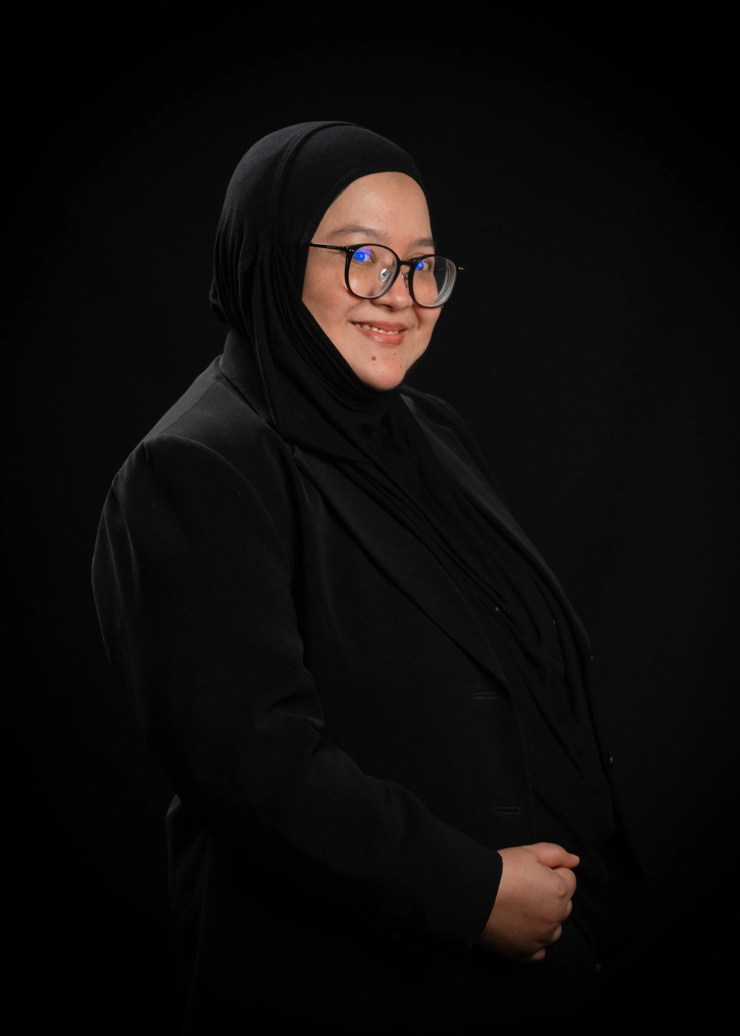 A woman wearing glasses and black hijab, smiling against a black background.