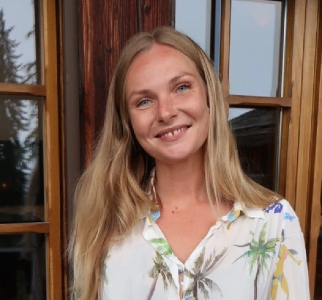 A woman with long blonde hair smiling in front of a wooden wall and glass windows.