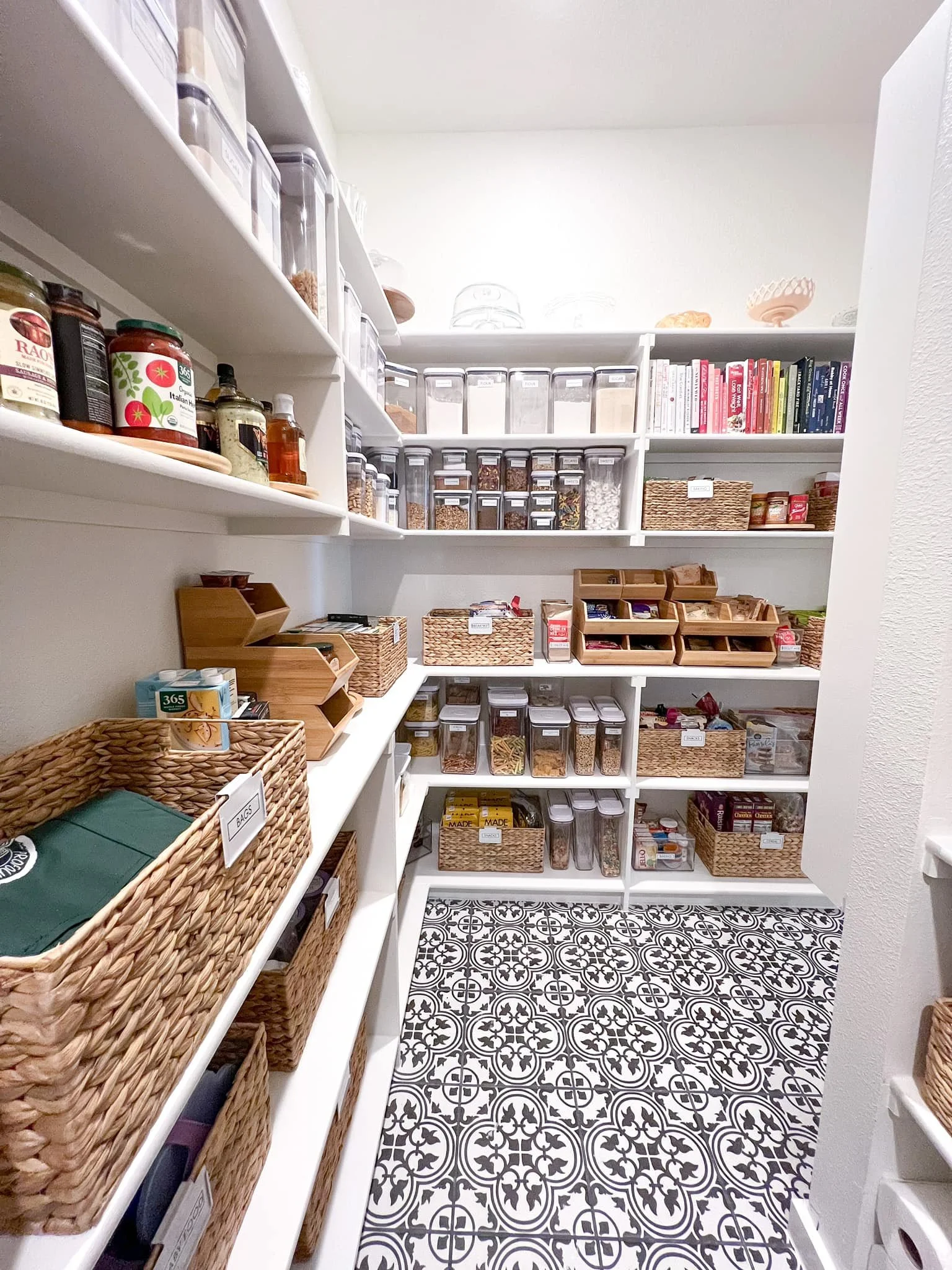 A well-organized pantry with white shelves filled with spices, grains, and food items in clear containers, along with wicker baskets and a decorative black and white patterned tile floor.