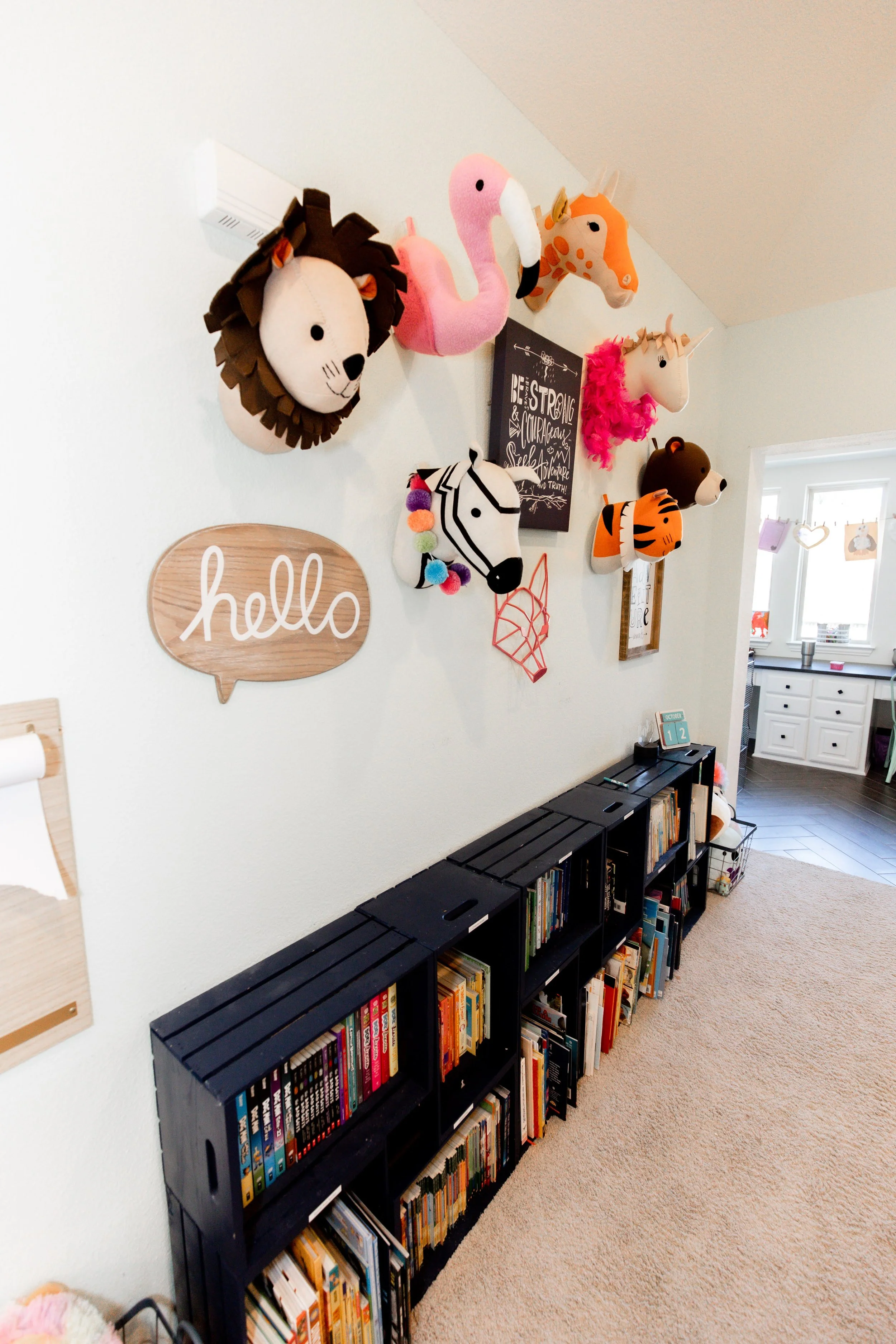 Decorative animal heads on a wall, including a lion, flamingo, giraffe, unicorn, zebra, bear, and tiger, with a wooden 'hello' sign below and black bookshelves filled with books on a beige carpeted floor.
