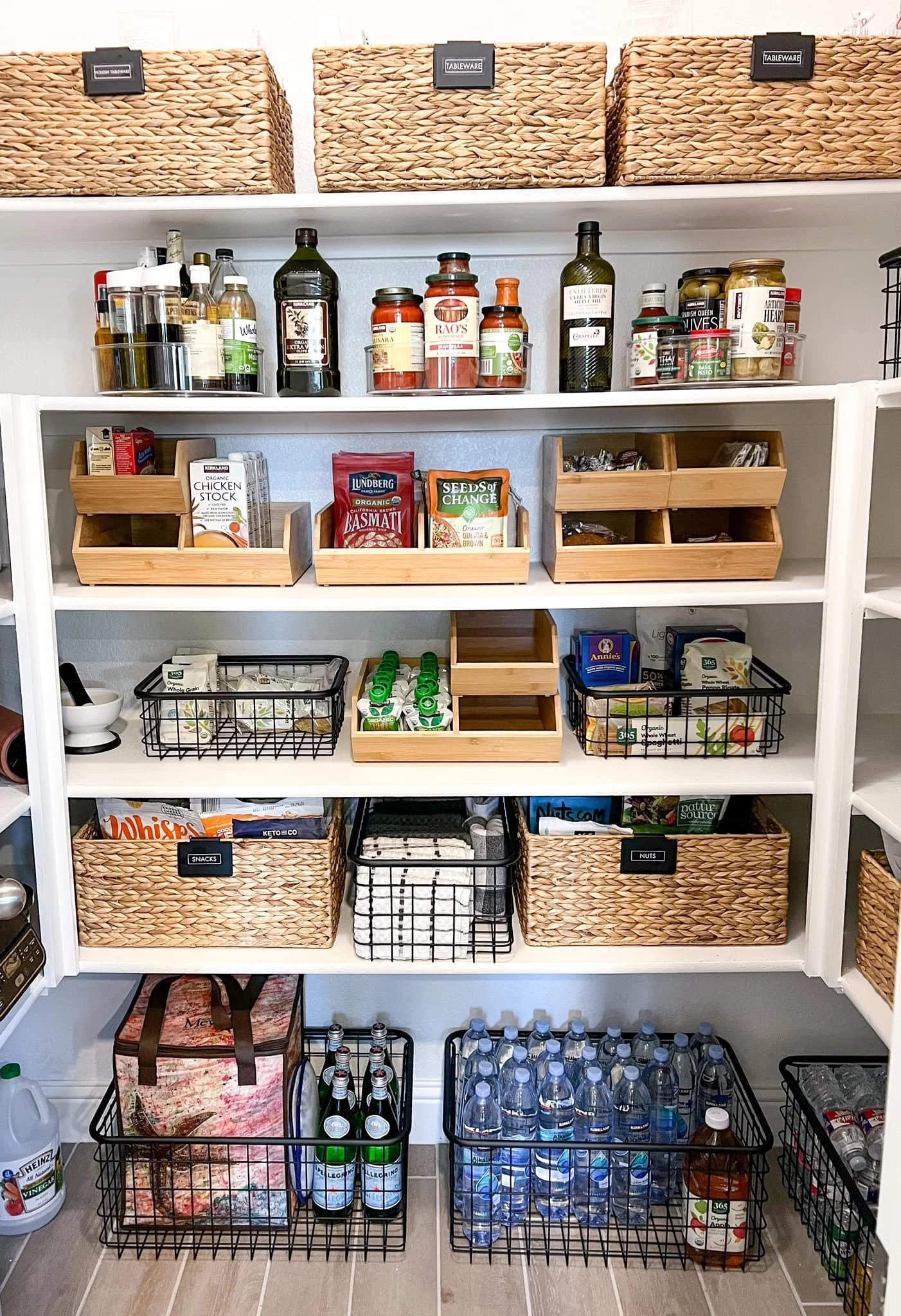 Pantry shelves with organized basket bins and various food and drink items, including bottled water, snacks, spices, and supplements.
