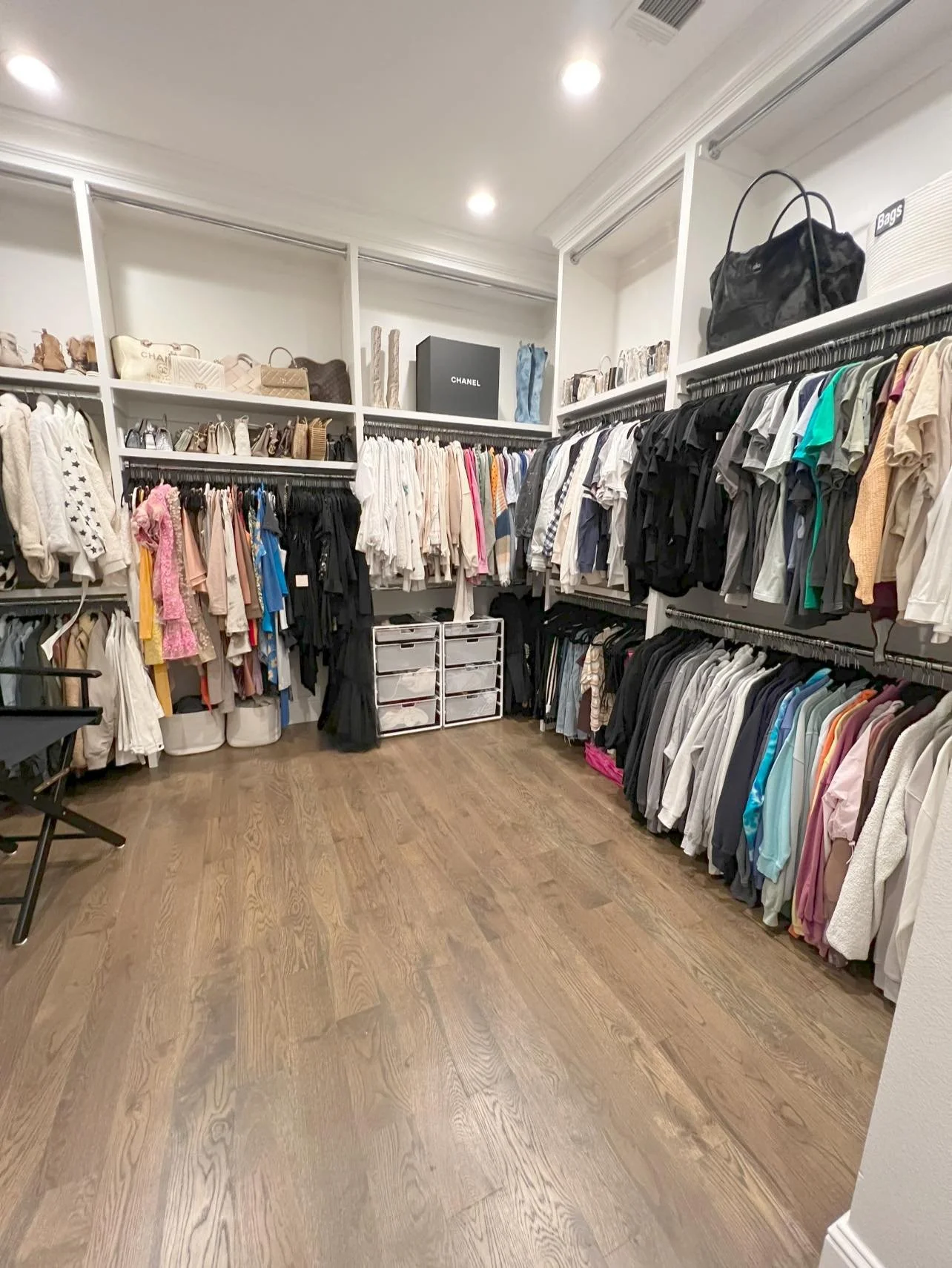 Closet with organized clothing on racks and shelves, handbags, and storage containers, with a wooden floor and recessed ceiling lights.