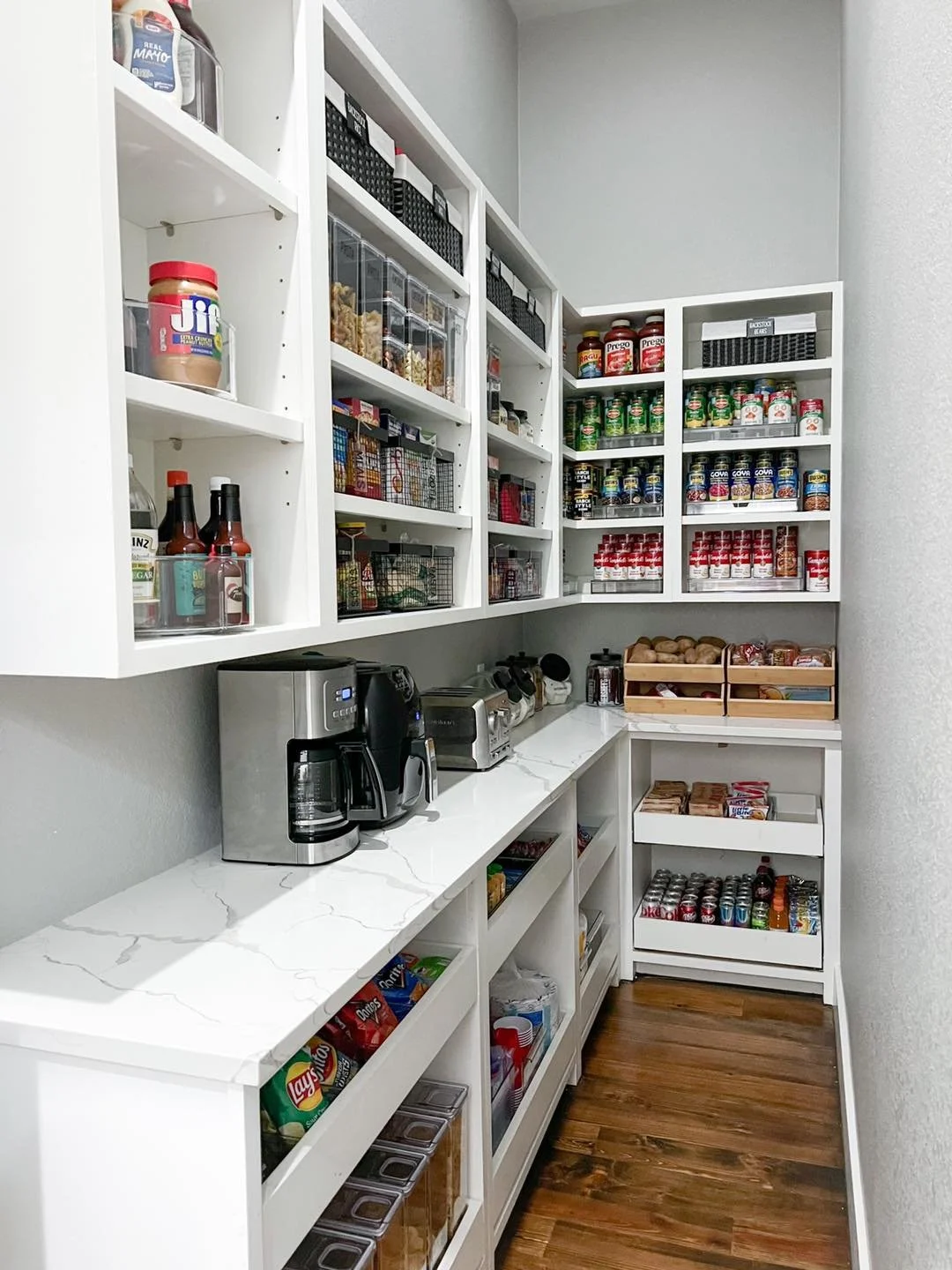 Pantry with white shelves filled with snacks, canned goods, spices, and appliances on white marble countertops.