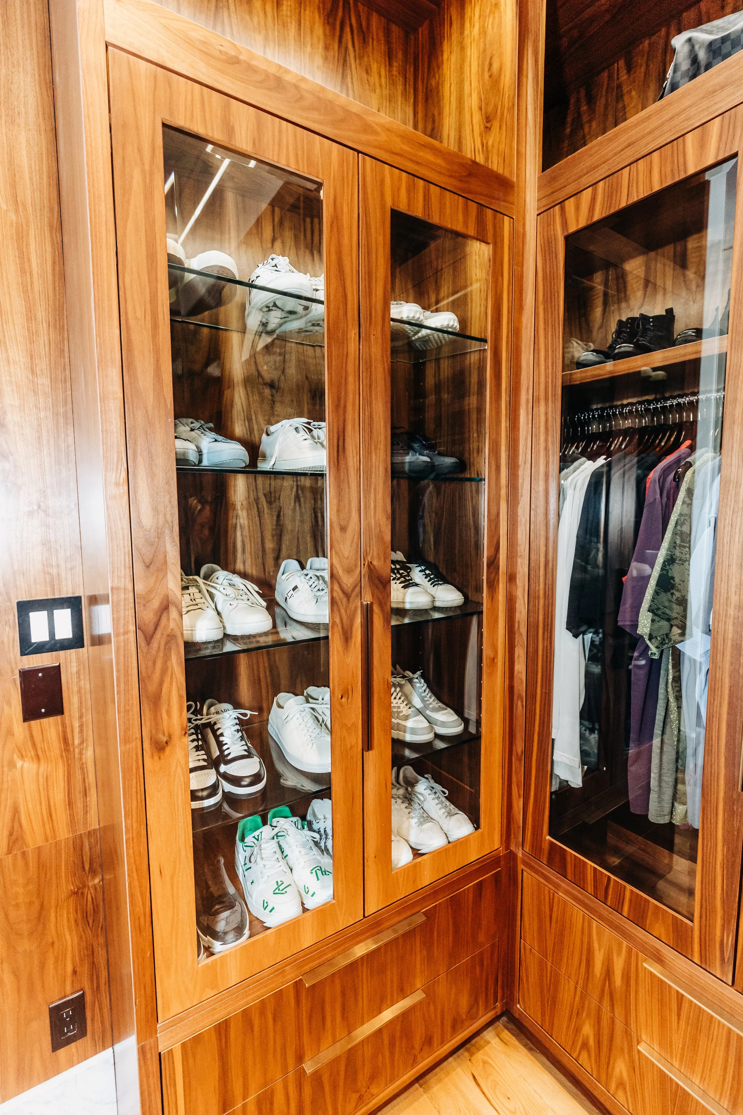 Built-in wooden closet with glass doors displaying white and black sneakers, and a section with hanging clothes.
