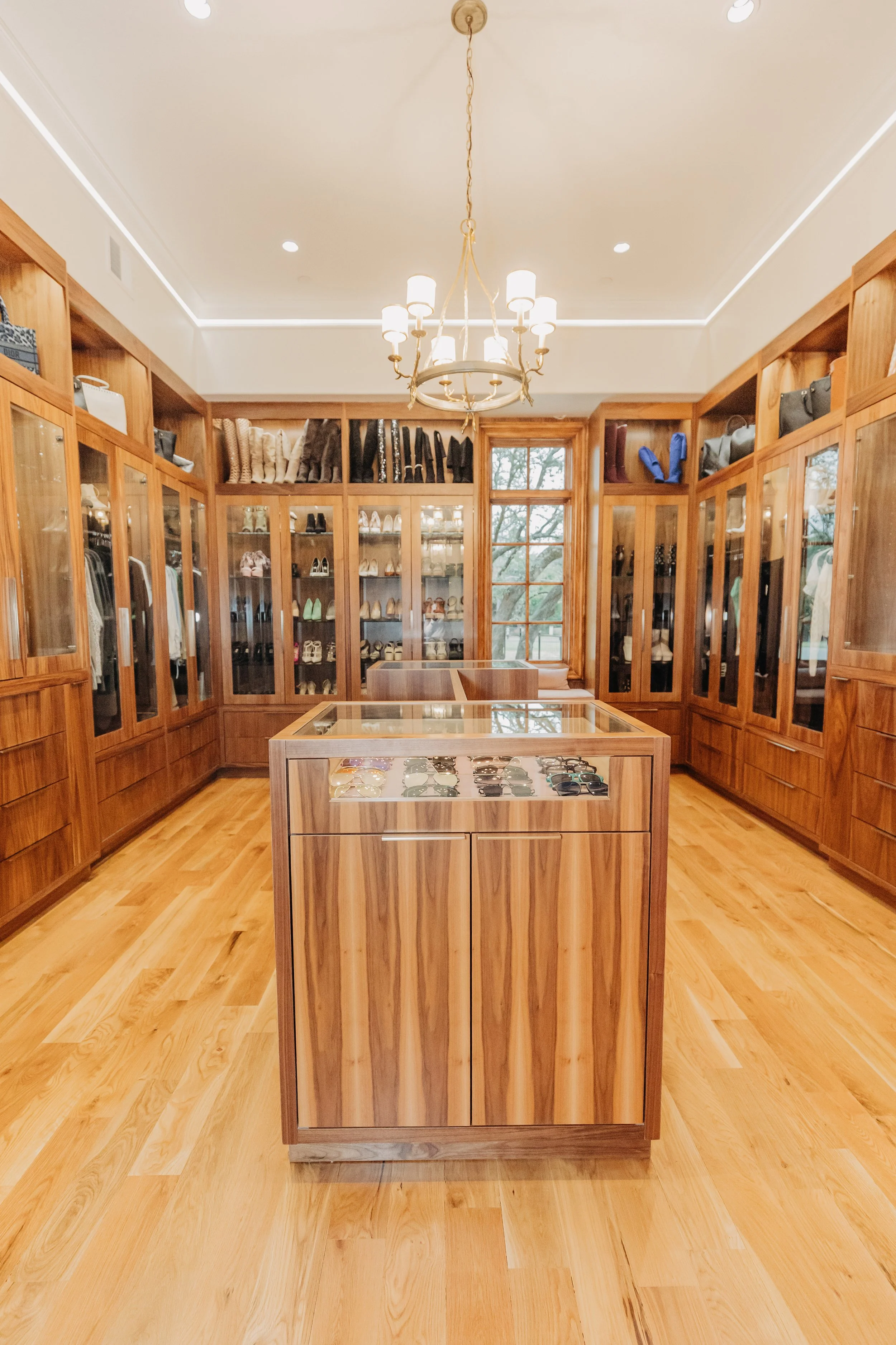A walk-in closet with wooden cabinets and a glass display island in the center, showcasing sunglasses. The room has a chandelier, recessed lighting, a large window, and shelves filled with shoes, handbags, and clothing.