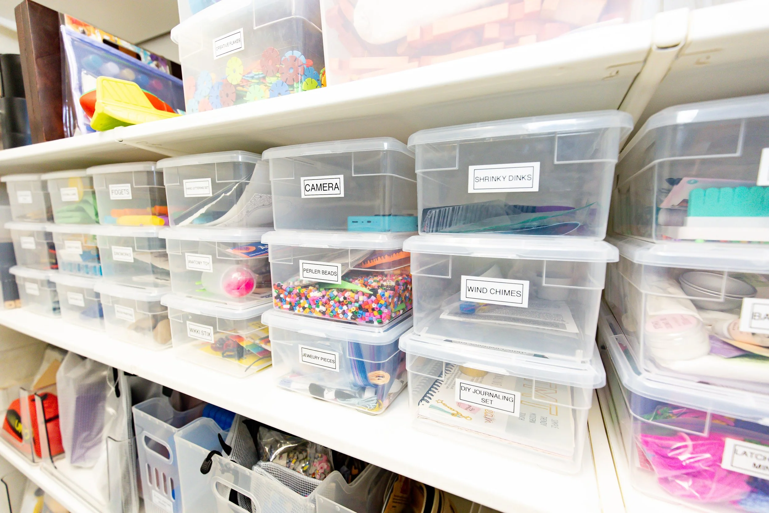 Clear plastic storage bins on a white shelf, labeled with items like 'Camera', 'Perler Beads', 'Wind Chimes', 'Shrinky Dinks', and others, containing craft supplies and small objects.