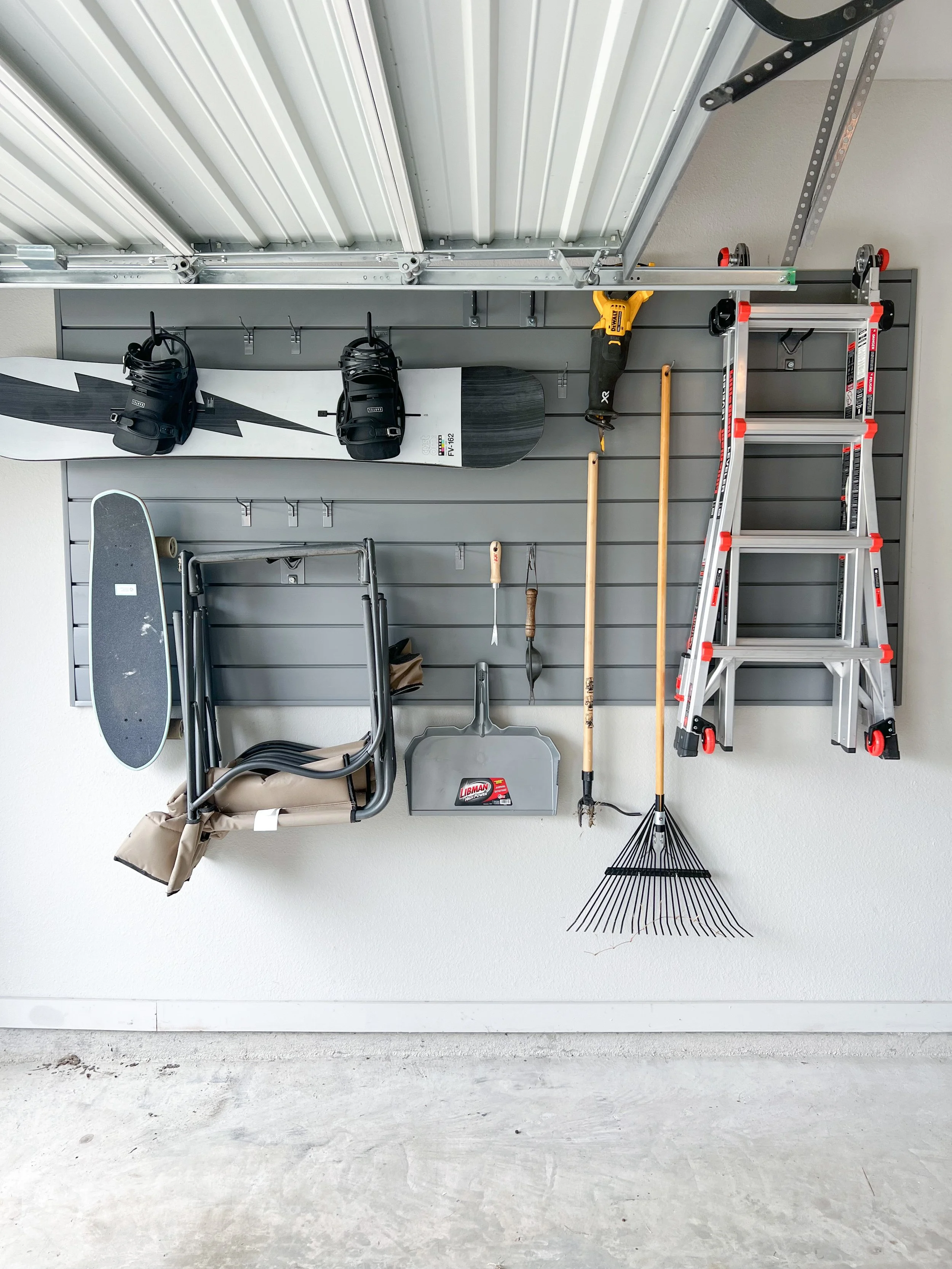 Garage wall with tools and equipment: skateboard, snowboard, two snowboard bindings, chainsaw, ladder, rake, shovel, folding chair, broom, and hooks on a slat wall with garage ceiling above.