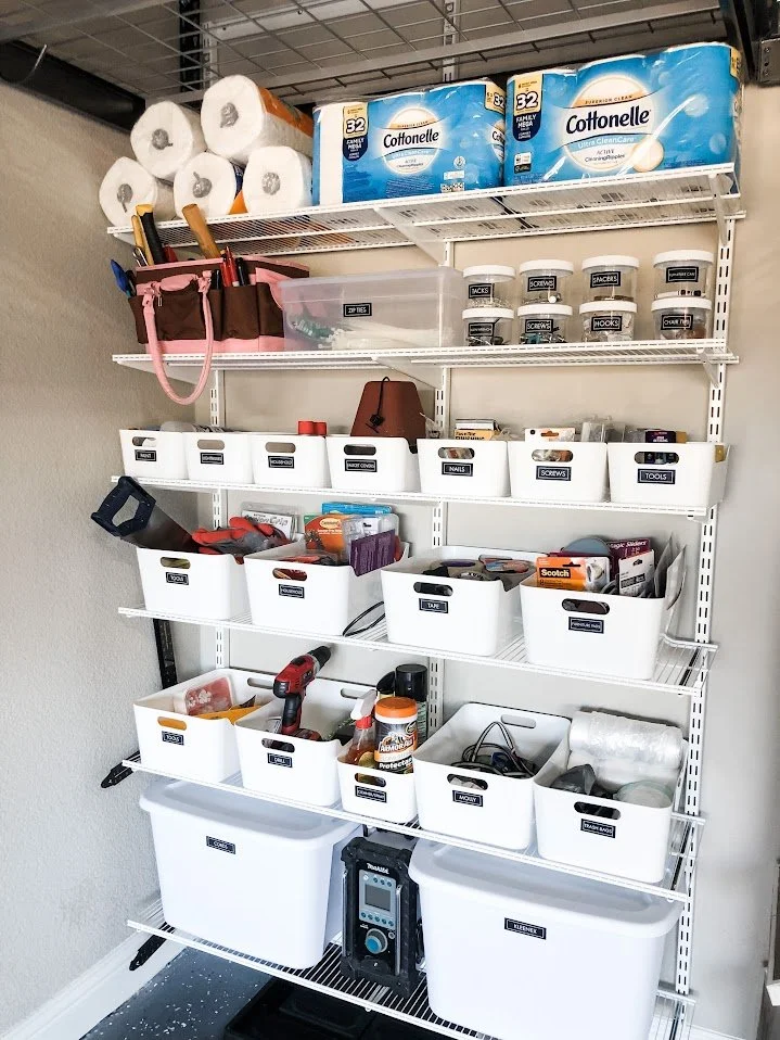 Garage storage shelving with various organized tools, hardware, and supplies, including paper towels, toilet paper, and storage bins labeled for different items.