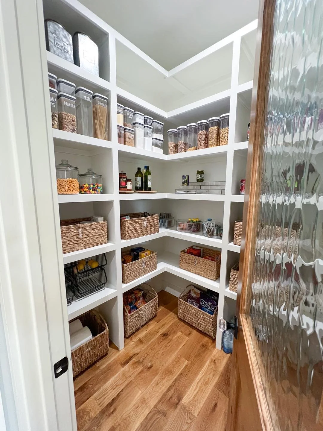 Pantry with white shelving filled with jars, baskets, and food items, and a wooden floor.