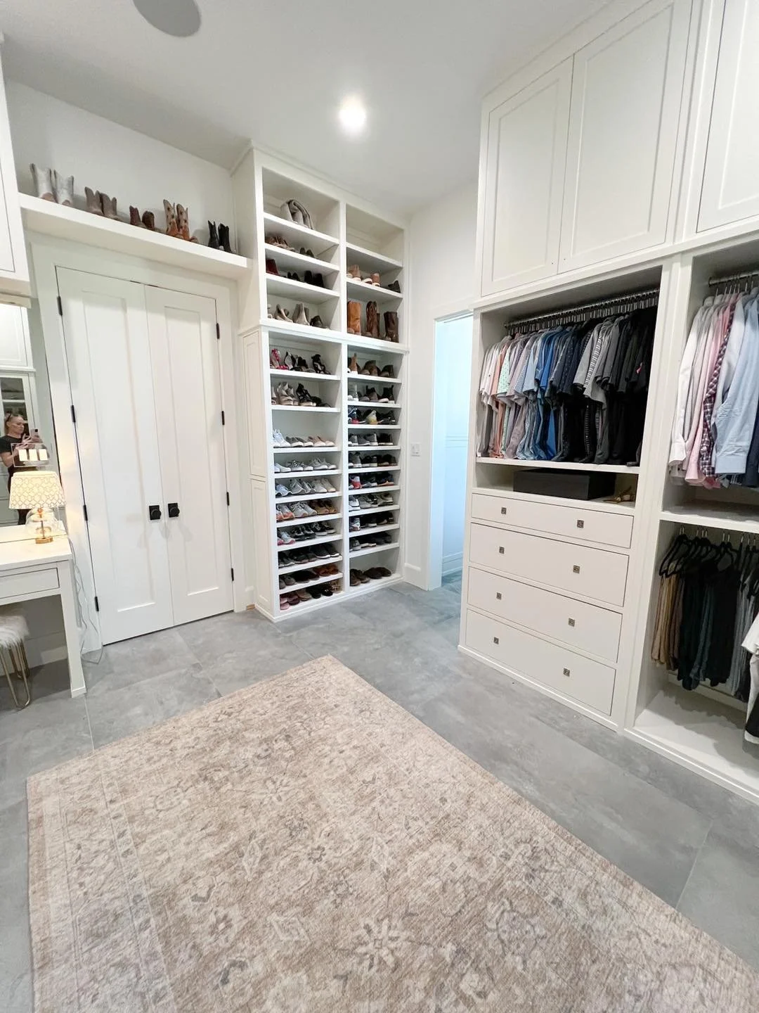 A spacious walk-in closet with white built-in shelves filled with shoes and folded clothes, a beige area rug on the grey tile floor, and a small white desk with a lamp.