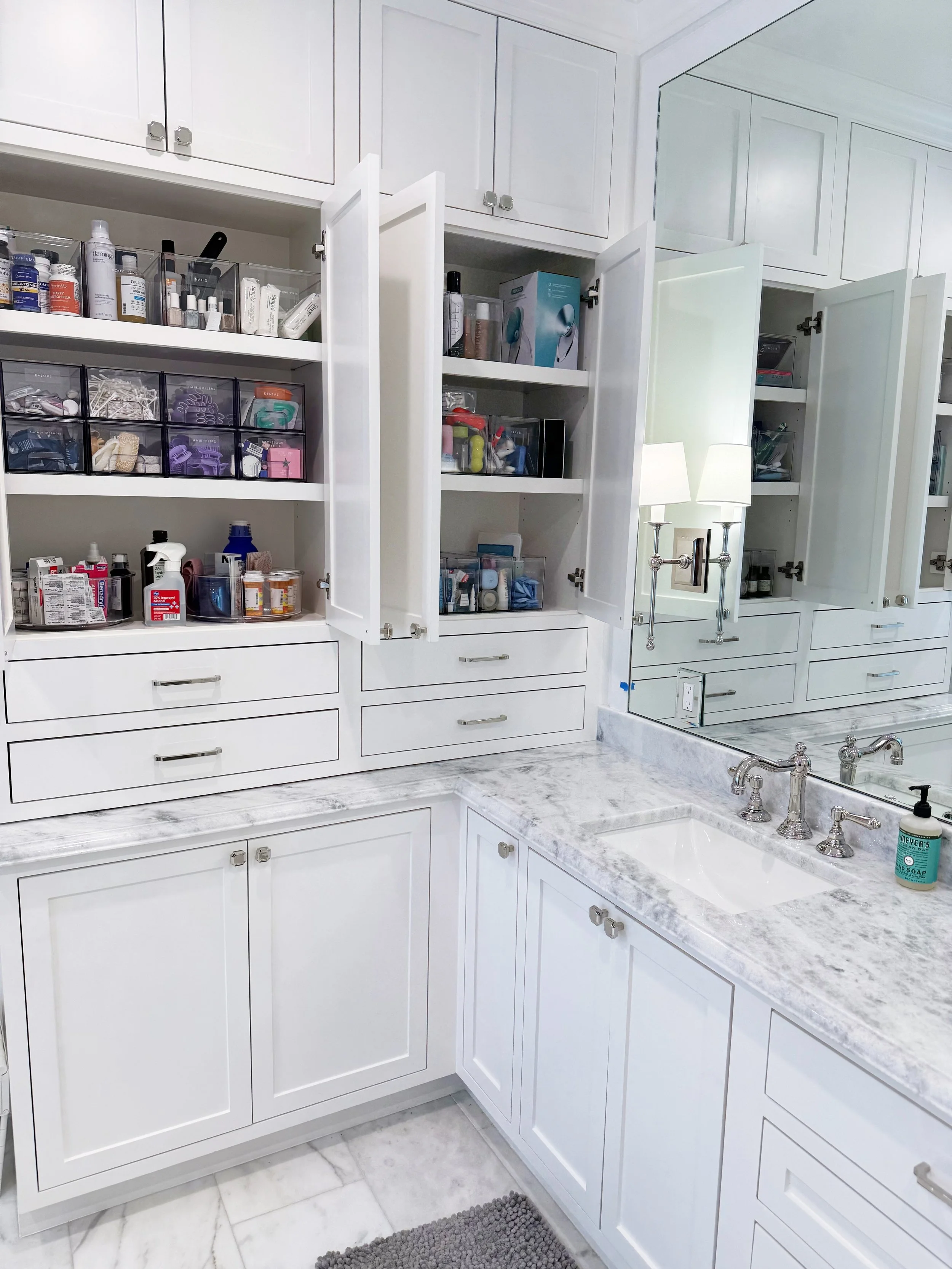 Bathroom with white cabinets, drawers, and marble countertop. Open cabinets reveal organized supplies and toiletries. A large mirror reflects part of the room, with wall-mounted sconce lights turned on.