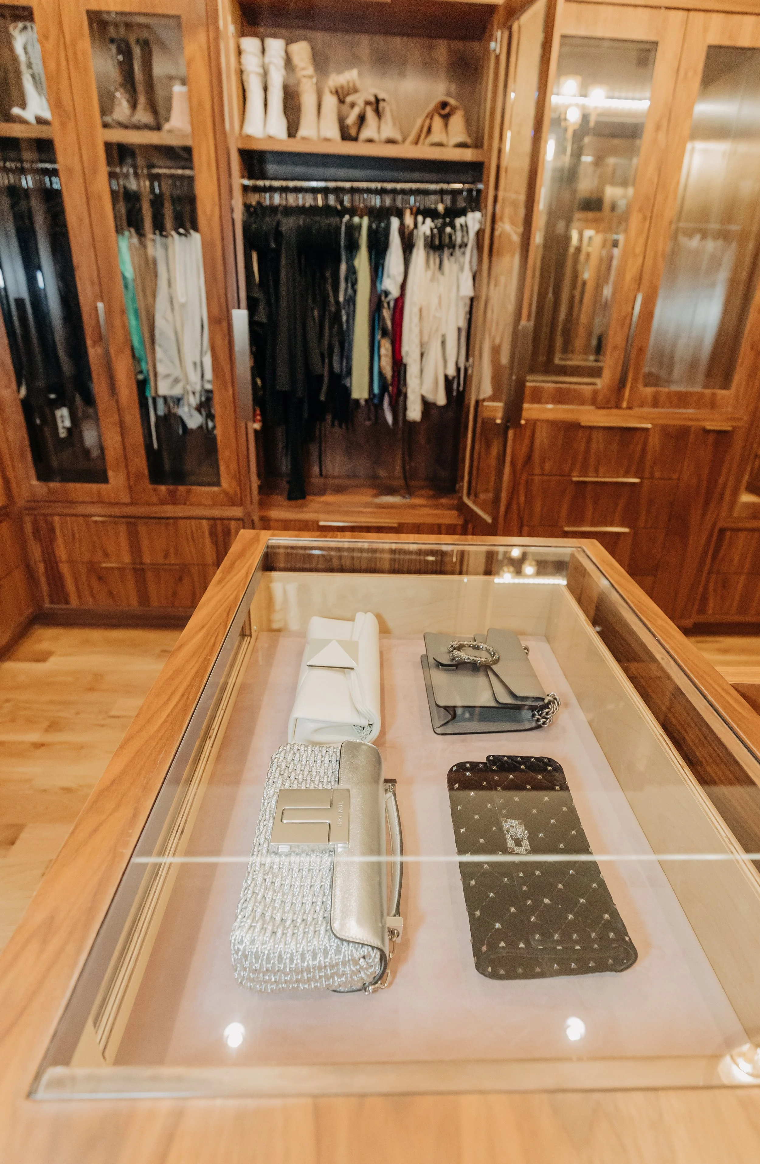 A glass display case with five designer handbags inside, set in a wooden room with closets filled with clothing and shoes in the background.