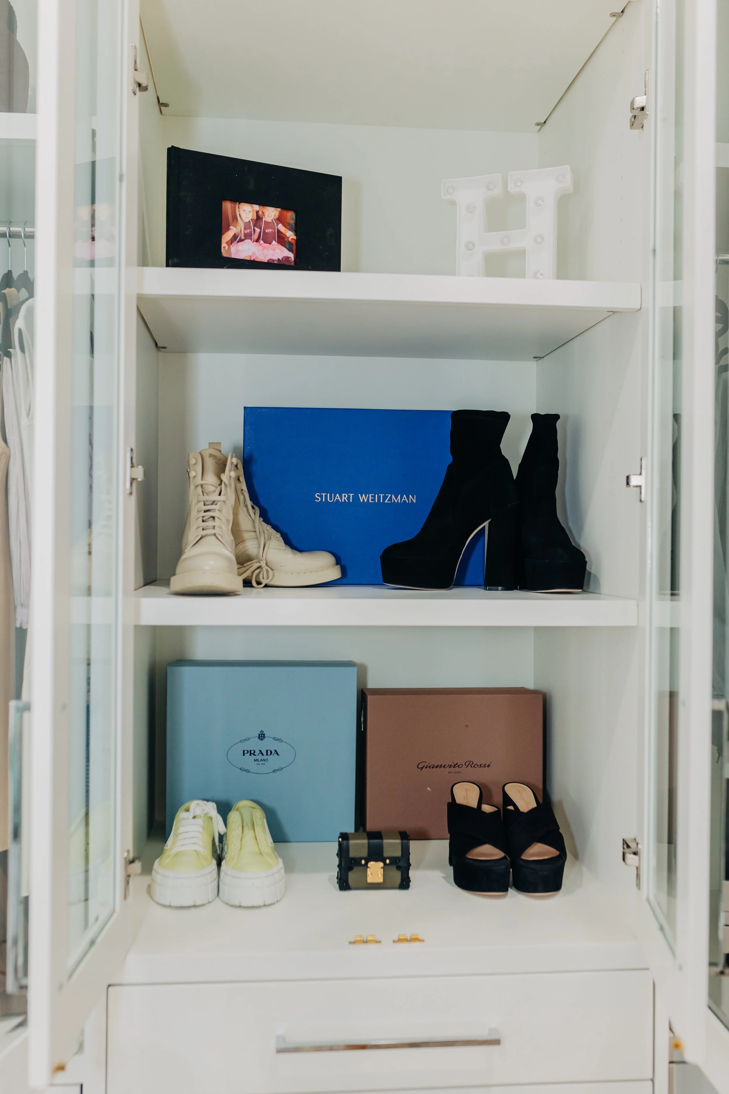A white display cabinet with two shelves holding various designer shoes and accessories, including a pair of cream-colored boots, black high-heeled boots, yellow sneakers, and black platform heels, along with designer boxes from Prada, Stuart Weitzma