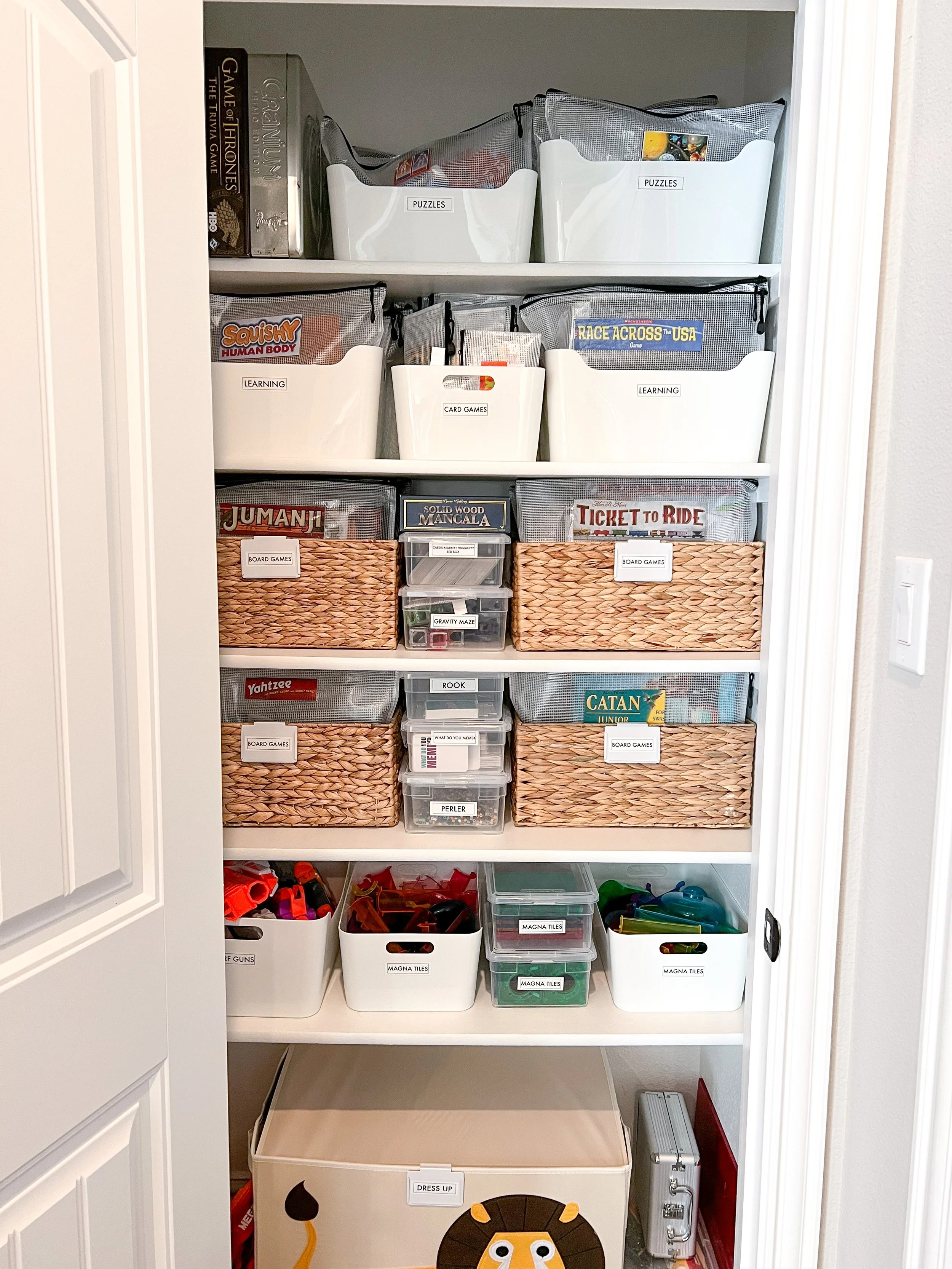 A neatly organized closet shelf with labeled storage bins containing board games, card games, puzzles, and toys, along with a large storage container at the bottom featuring a lion face.