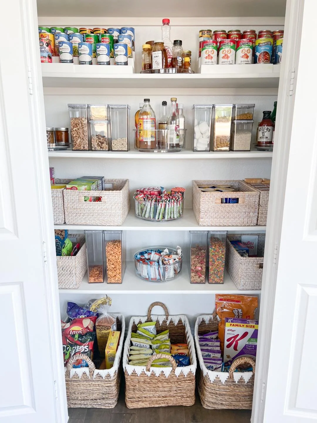 A pantry with four shelves filled with canned goods, jars, and boxed items, and three baskets at the bottom containing snack packages.