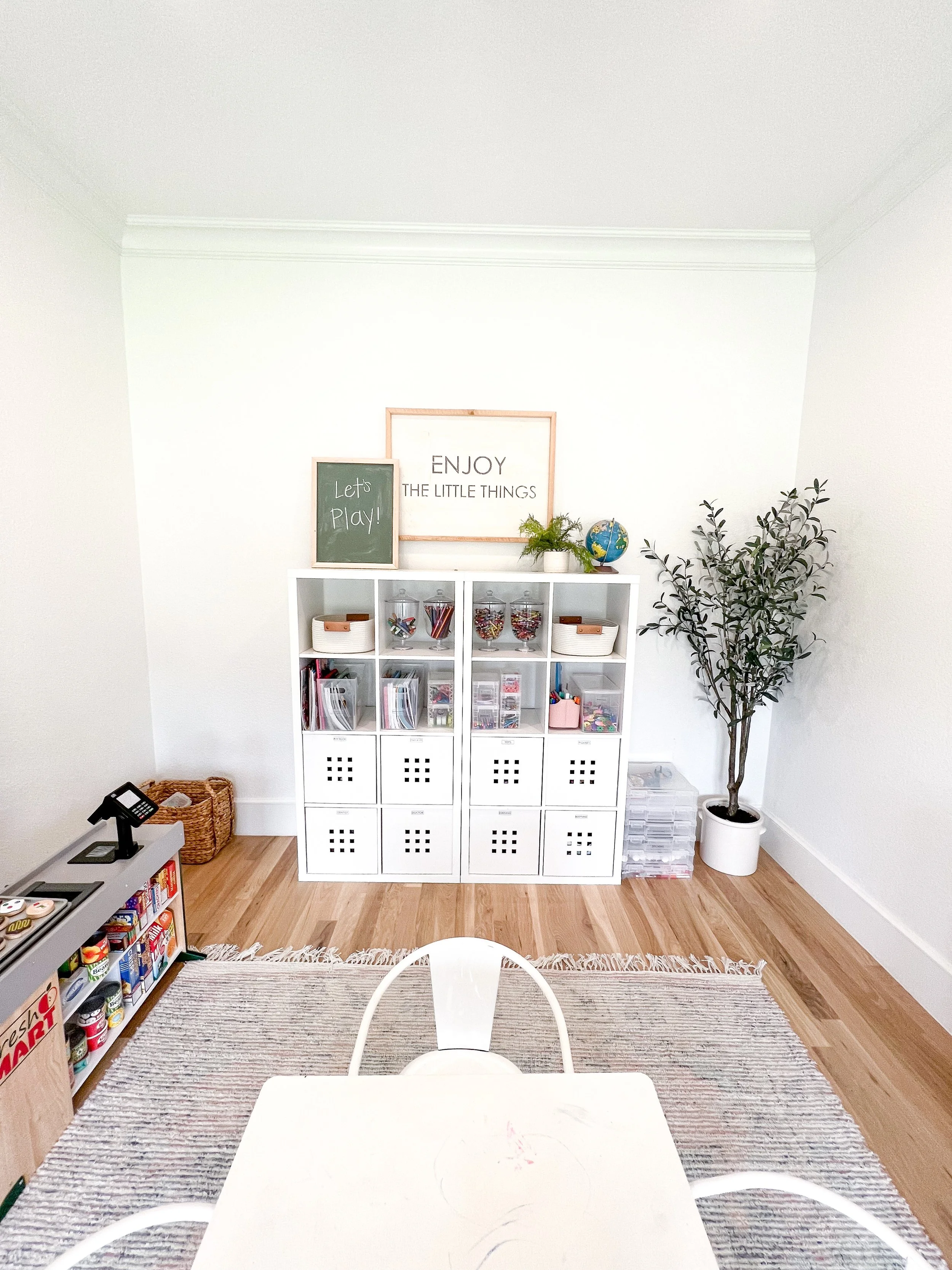 A children's playroom with white shelving, a small table and chairs, toy storage, and wall decor.
