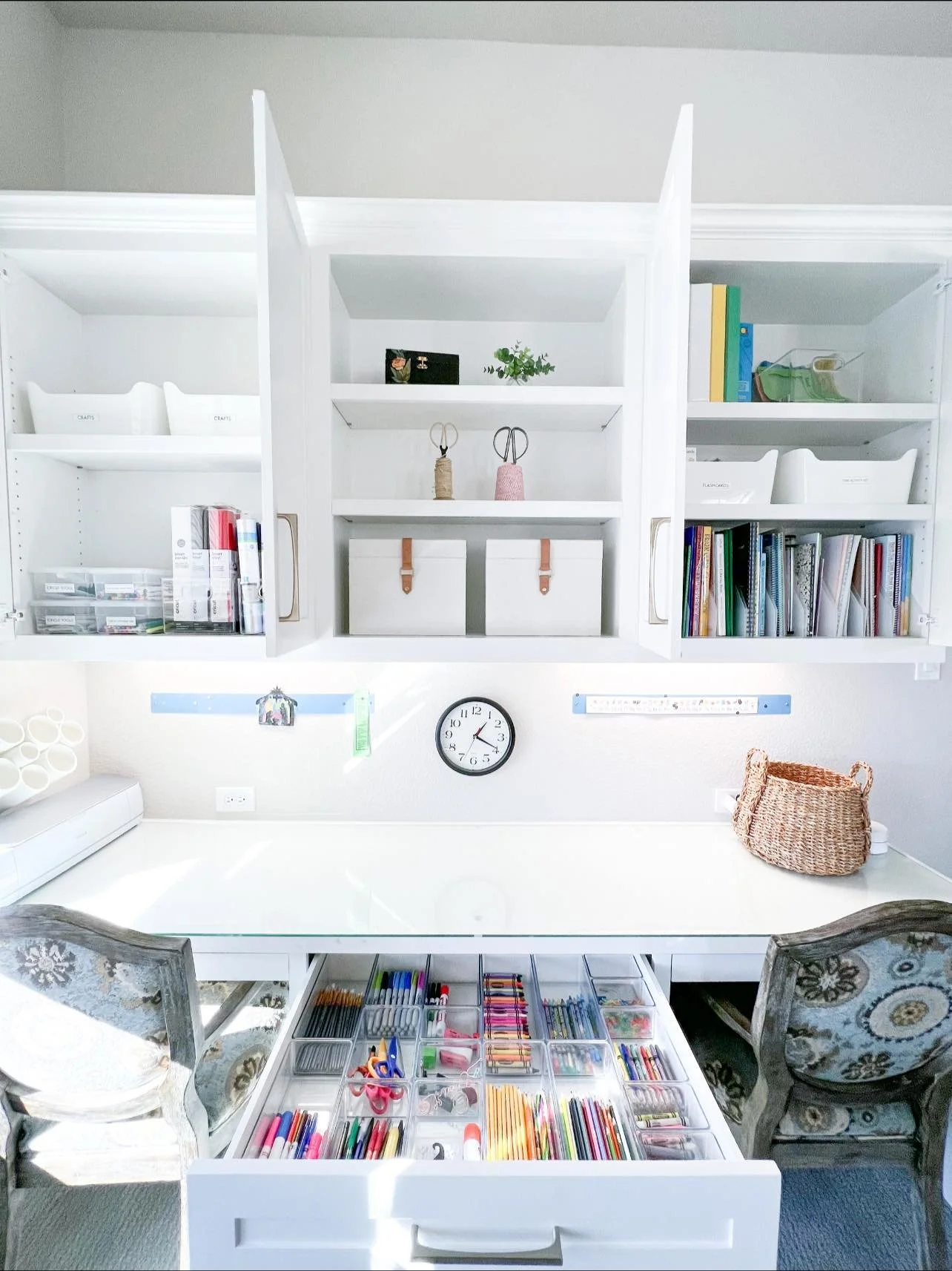 A well-organized craft room with a white work table, two patterned chairs, a wall clock, and white wall-mounted cabinets filled with books, binders, and craft supplies. The drawer in the table displays an assortment of colored markers, scissors, and 