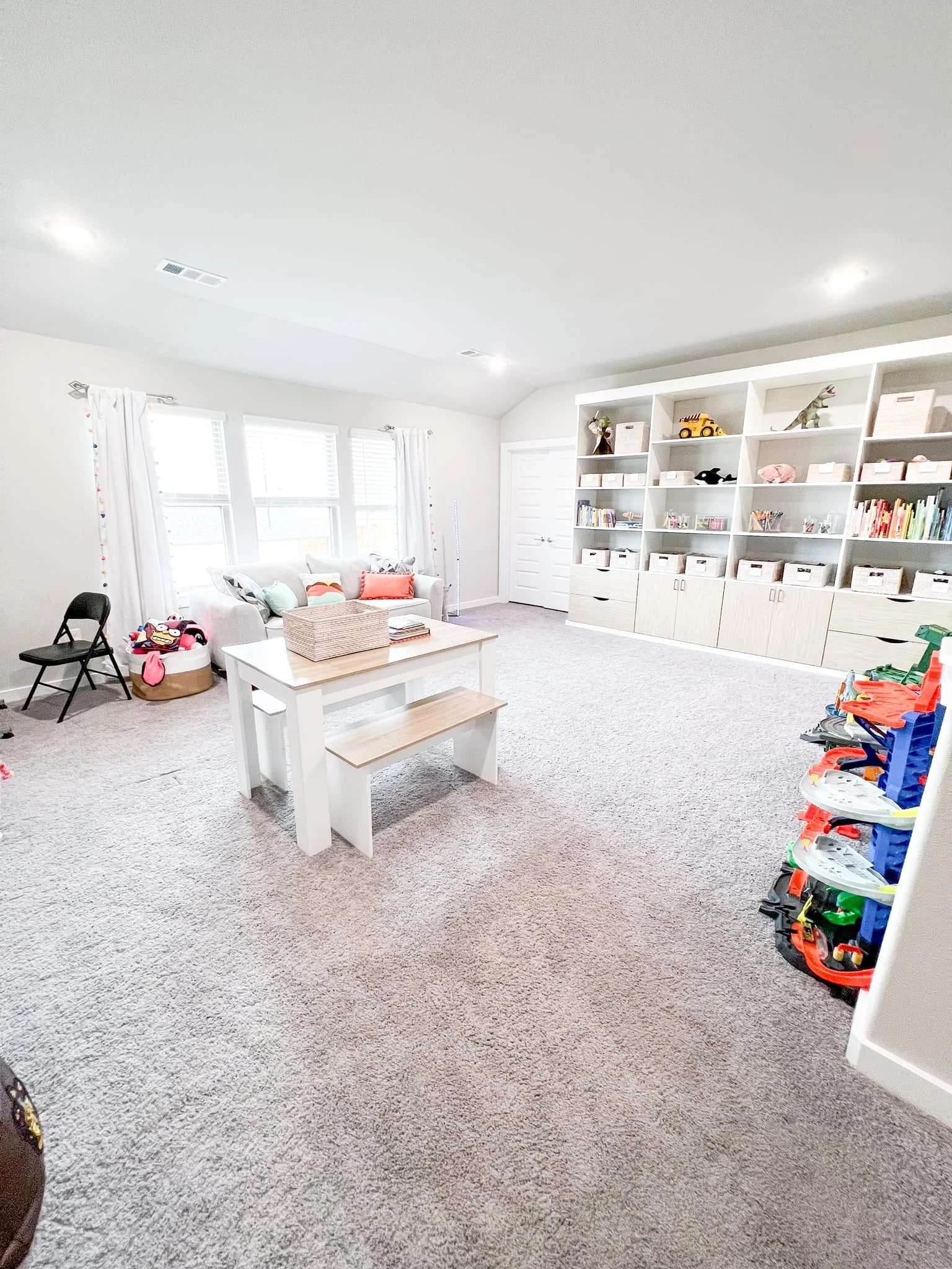 A brightly lit playroom with white walls, beige carpet, white furniture, storage shelves filled with toys and books, and a window with white curtains.