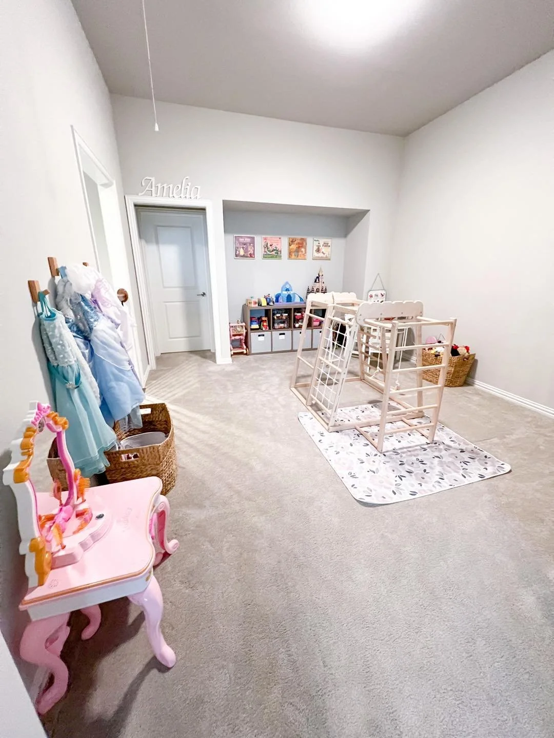 Empty children's playroom with a pink vanity table, dress rack, and toy storage, with a partially assembled crib and a white rug, on beige carpeted floor.