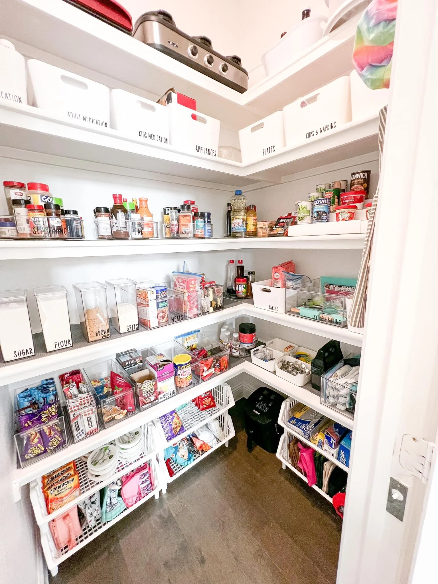 Organized pantry with labeled containers, spices, snacks, baking supplies, and kitchen appliances on shelves and in bins.