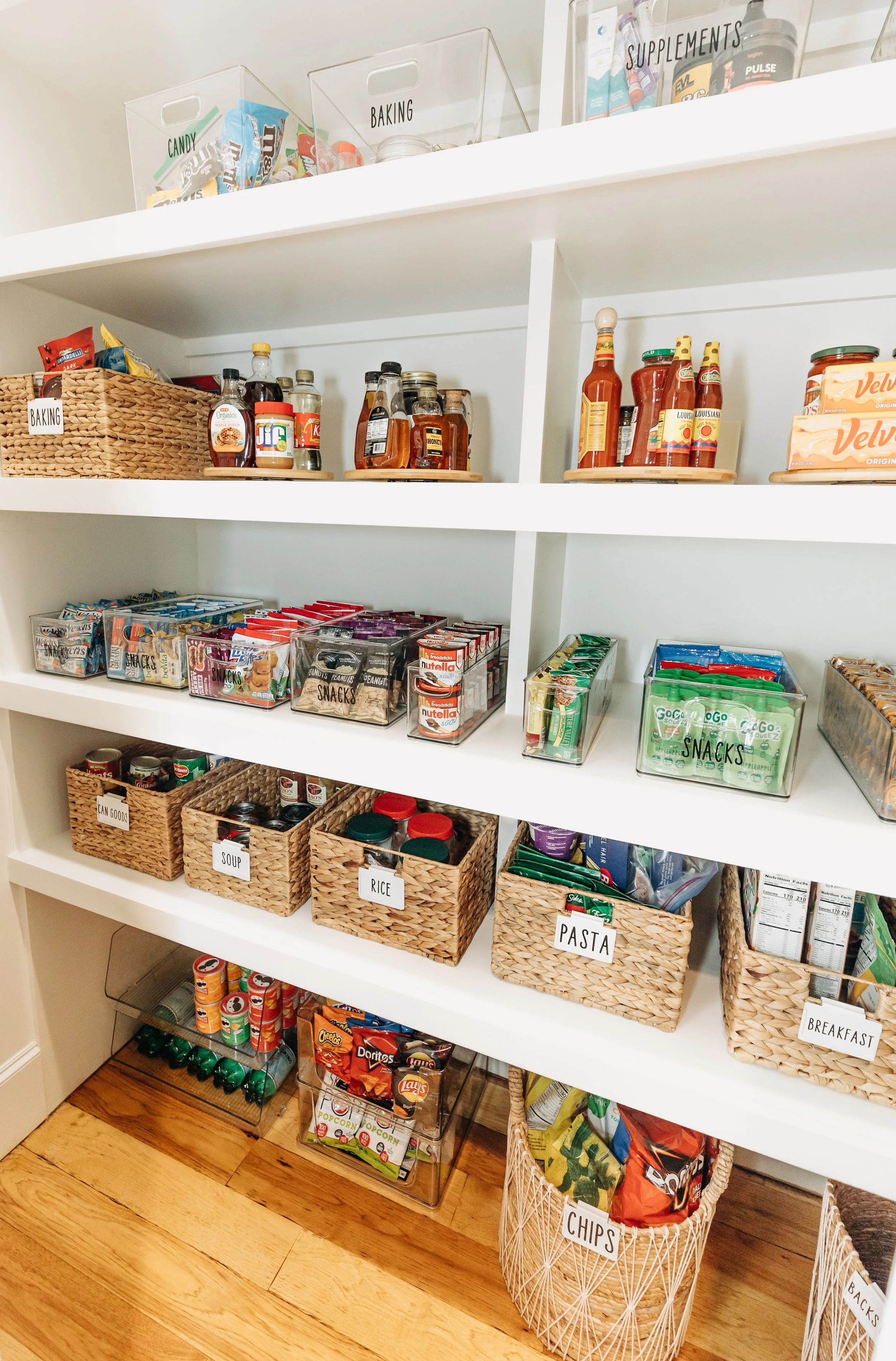 Pantry shelf with labeled baskets containing food and snack items, including cans, rice, pasta, soup, chips, and organized jars of condiments and supplements.