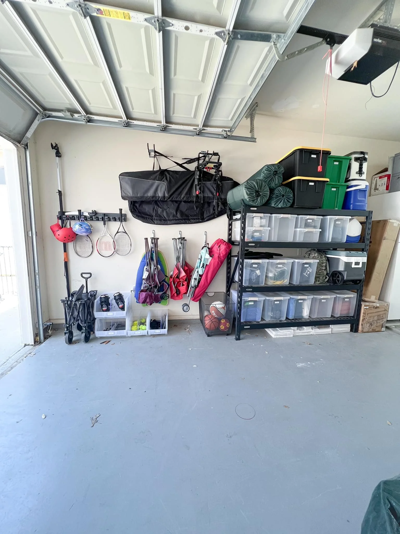 A garage wall with organized sports equipment and storage containers, including tennis rackets, balls, sports gear, and various bins on metal shelves.