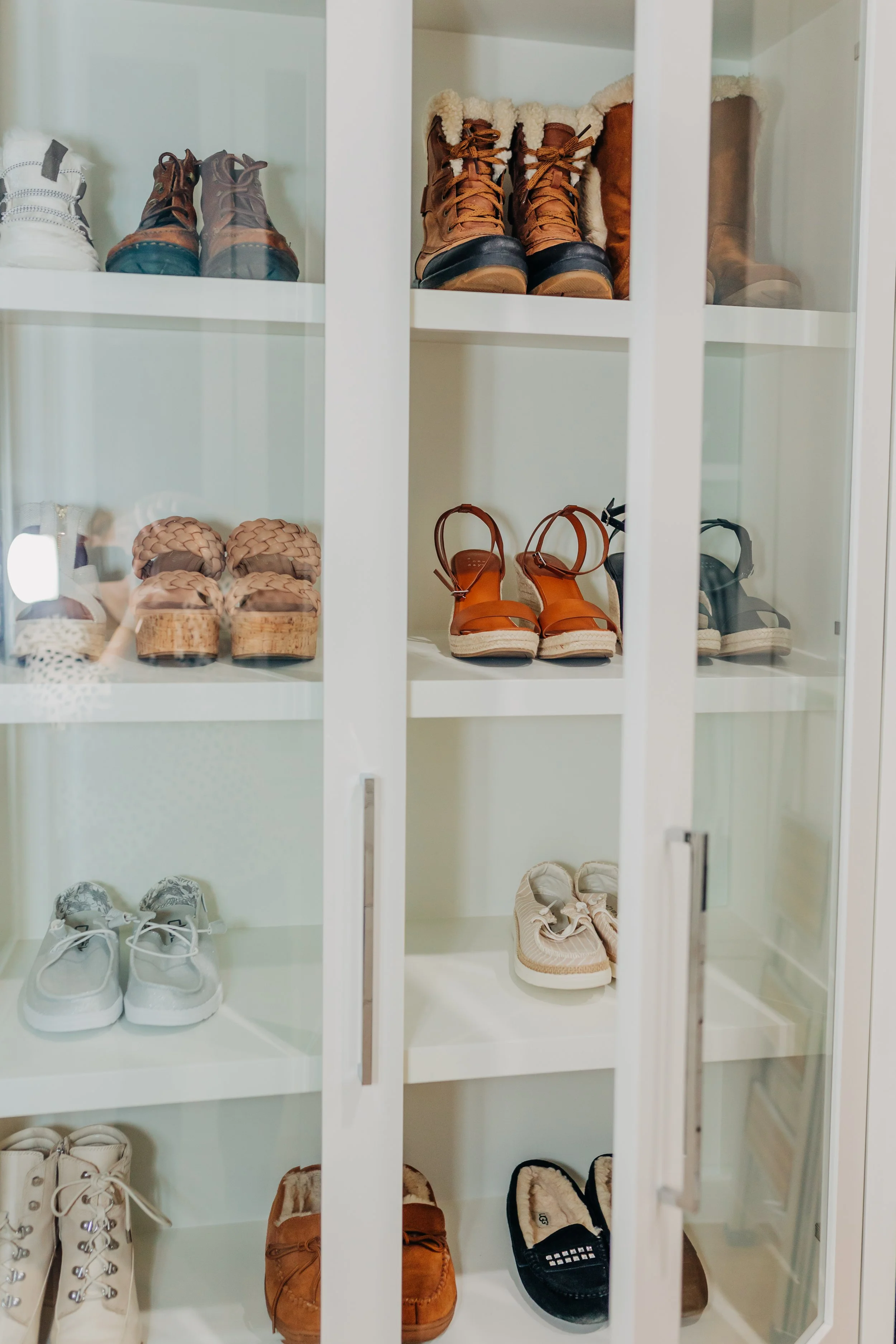 A glass-front white shoe cabinet filled with various pairs of shoes and boots on multiple shelves.