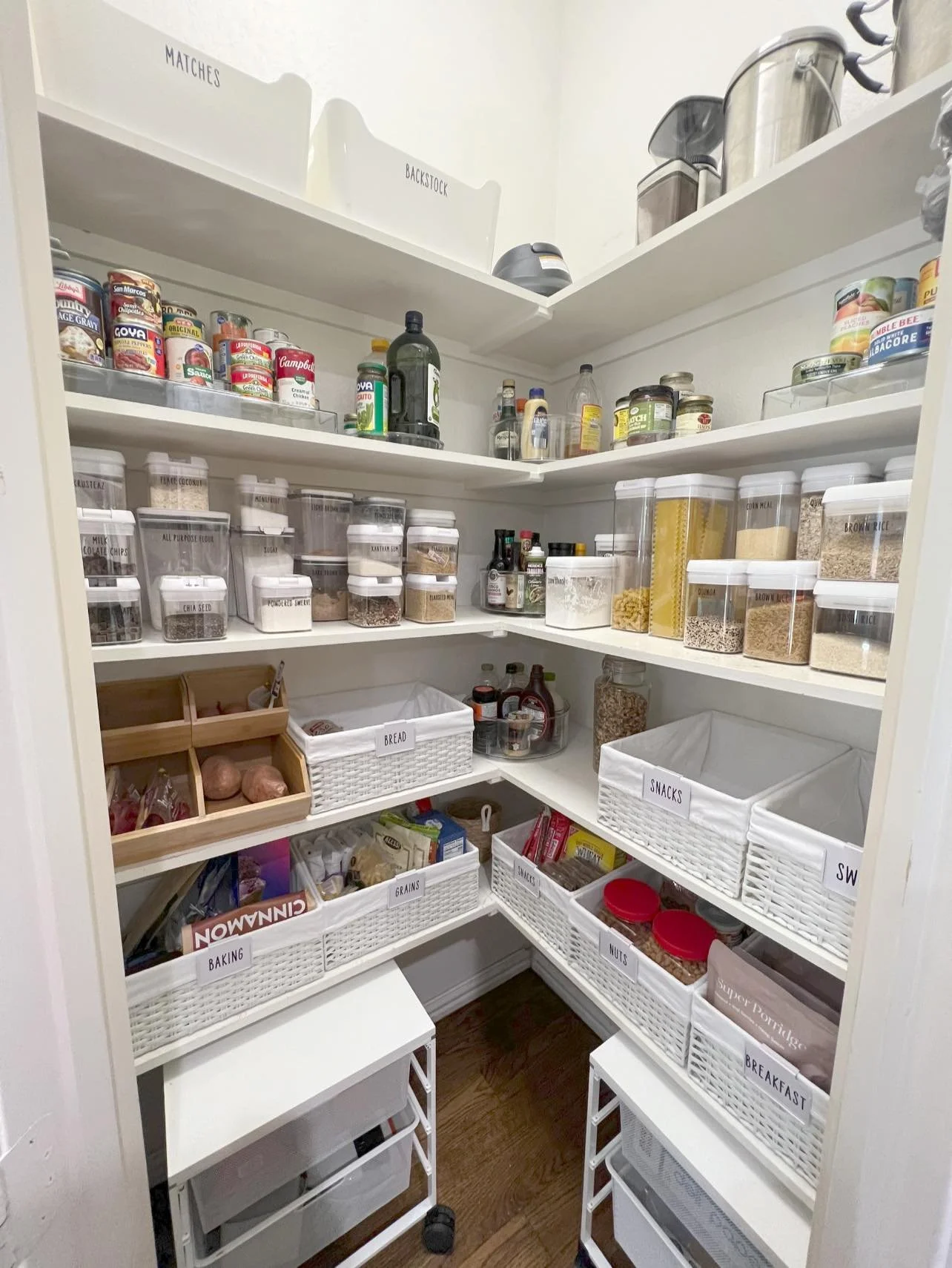 A well-organized pantry with labeled white baskets containing grains, snacks, nuts, breakfast items, and baking supplies. Shelves above hold canned goods, bottles, and jars, with baking tools on the top shelf.