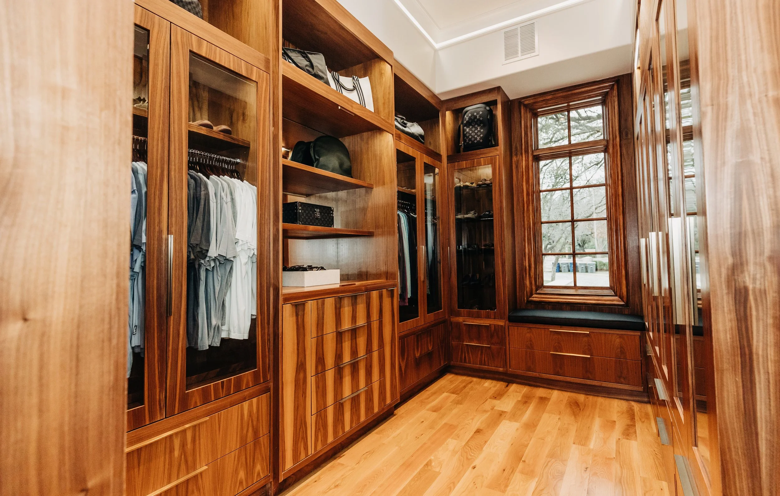 Wood-paneled walk-in closet with built-in shelves, glass-front cabinets, and a window seat with a black cushion.