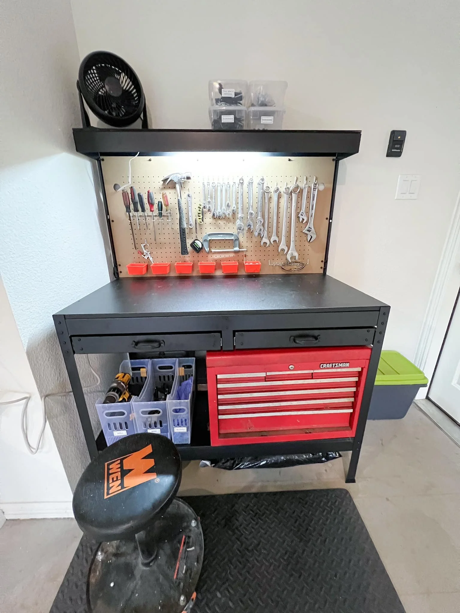 A black workbench with a red tool chest, pegboard with various hand tools, and plastic bins on top. There is a fan on the upper left corner, a stool with WEN logo, and a green storage bin on the right.