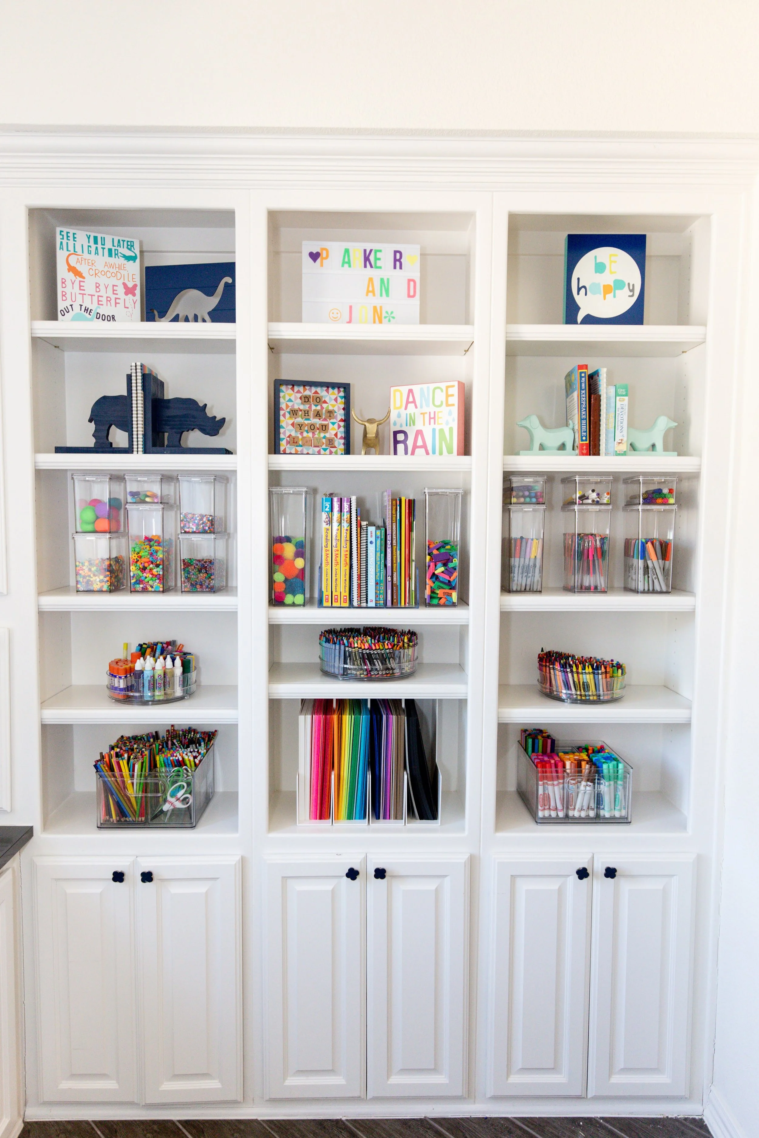 White bookshelf filled with colorful books, arts and crafts supplies, and decorative items including signs with playful designs and messages, resembling a child's creative space.