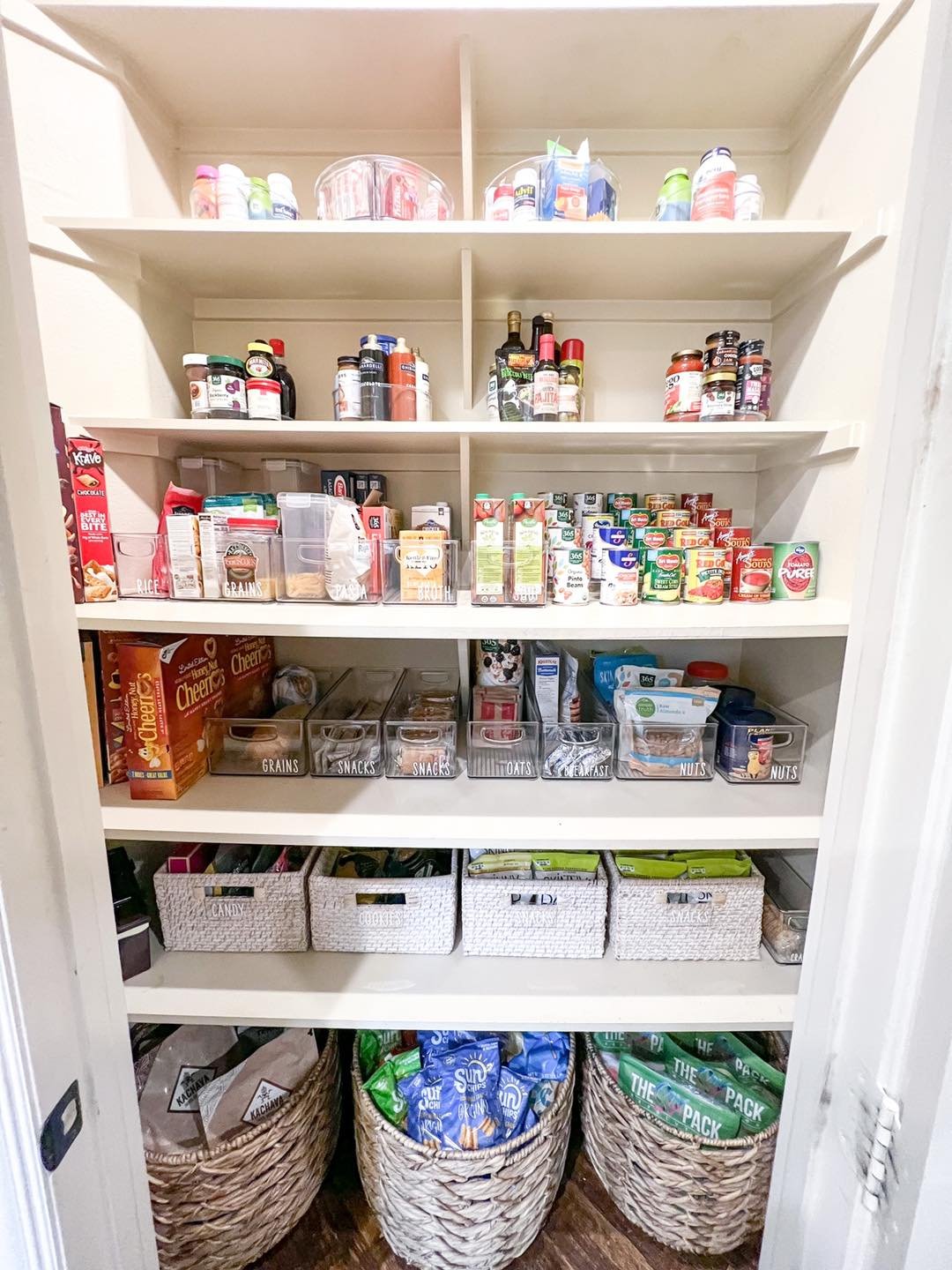 Organized pantry with five white shelves filled with various food and supplement jars, cans, and boxes, along with four woven baskets containing snacks and packaged foods on a wooden floor.