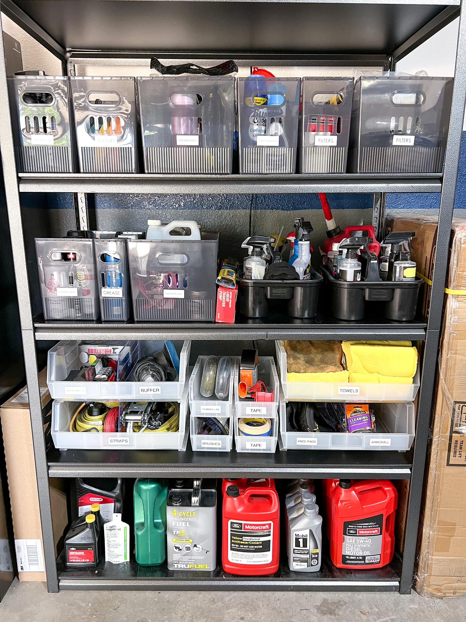 Metal shelving unit containing automotive and cleaning supplies, organized in labeled bins. Top shelf has filters and car wash supplies; middle has spray bottles and lubricants; lower shelves hold tools, tape, towels, and vehicle fluids.