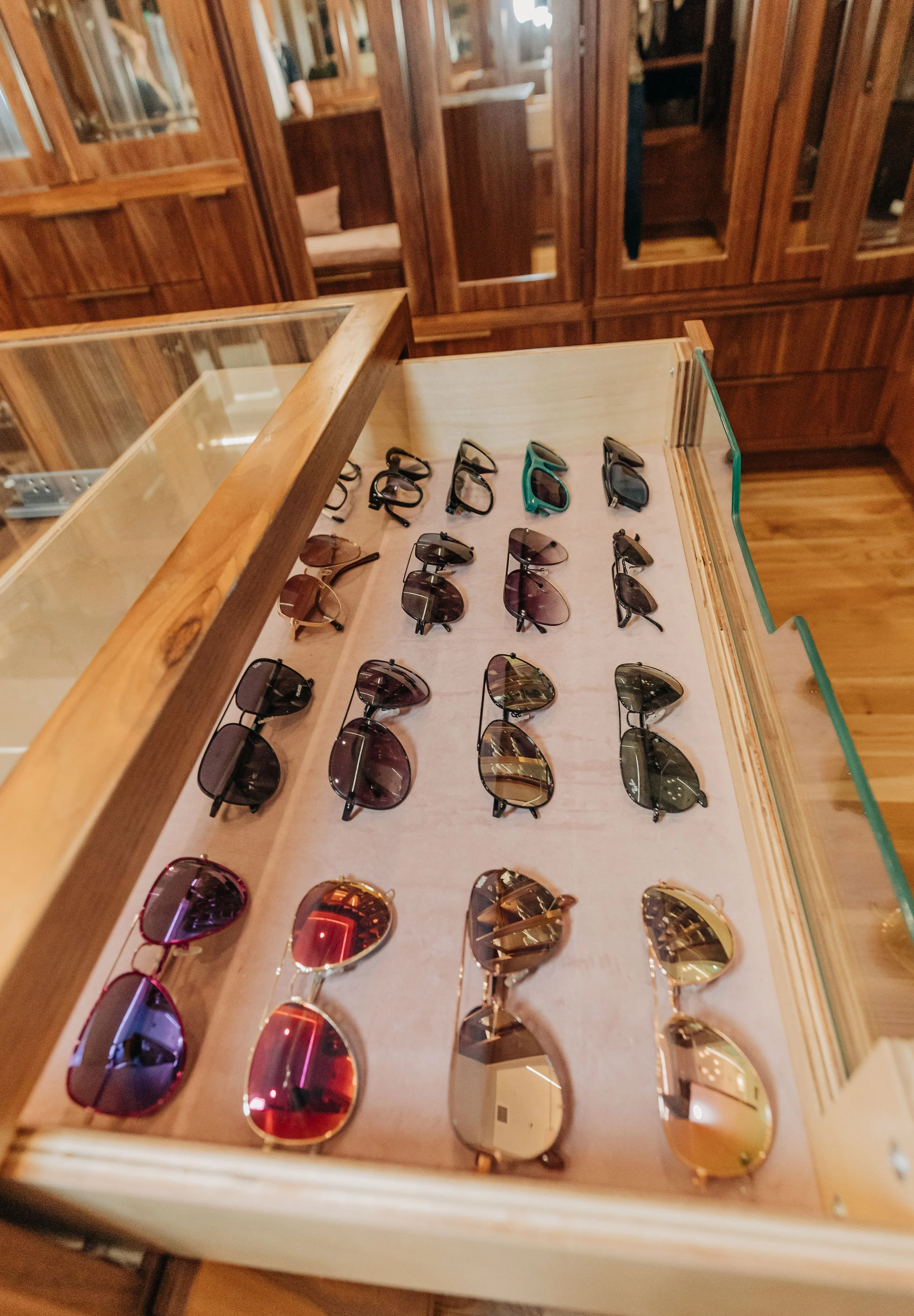 Display case with multiple pairs of sunglasses in various styles and colors inside a store with wooden shelves.