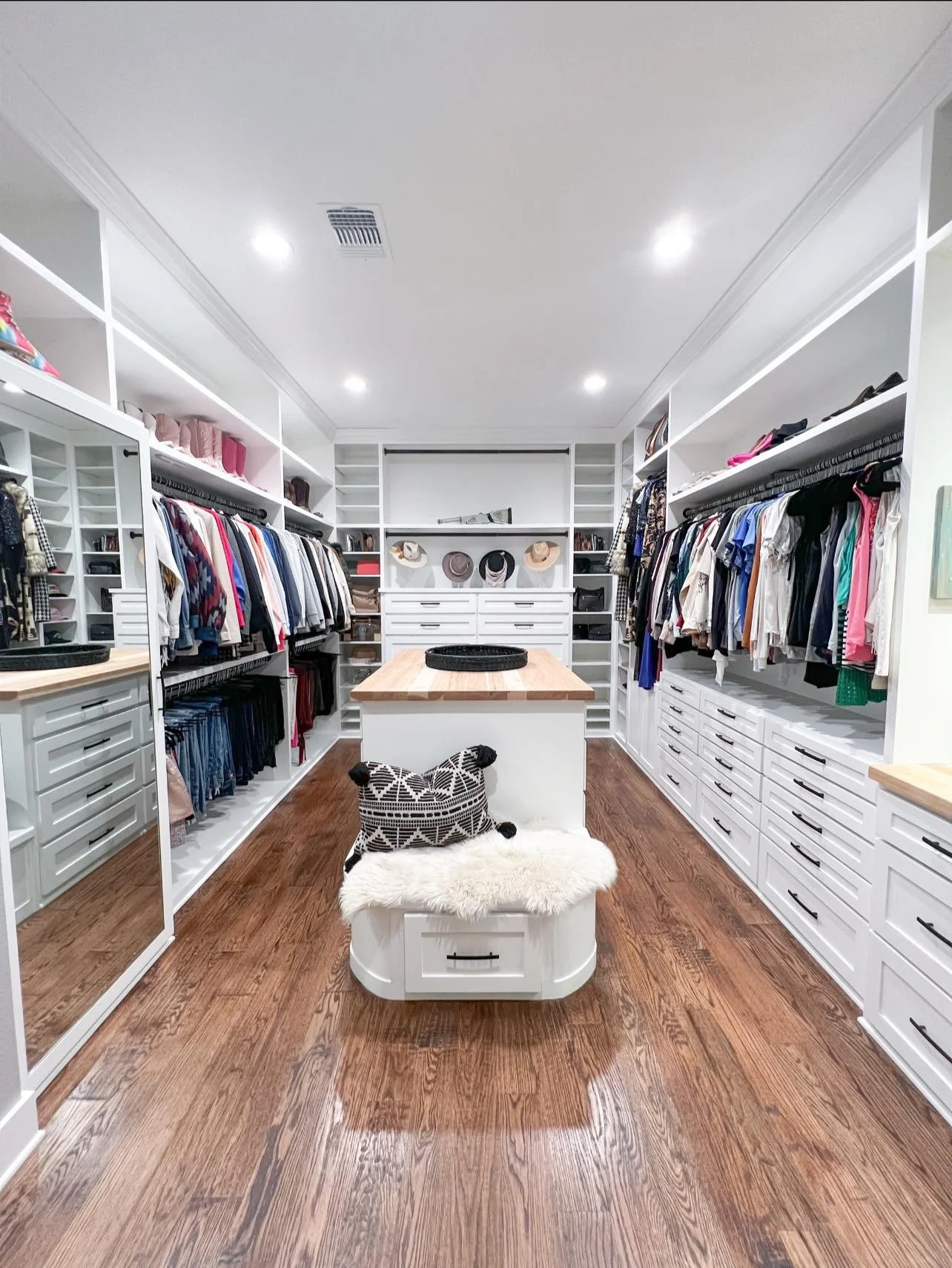 A walk-in closet with white shelves and drawers filled with clothes, shoes, and accessories. A central island with a wooden top, decorative hats, and a black tray. A bench with a furry white cushion and a black and white pouch in the foreground. Hard