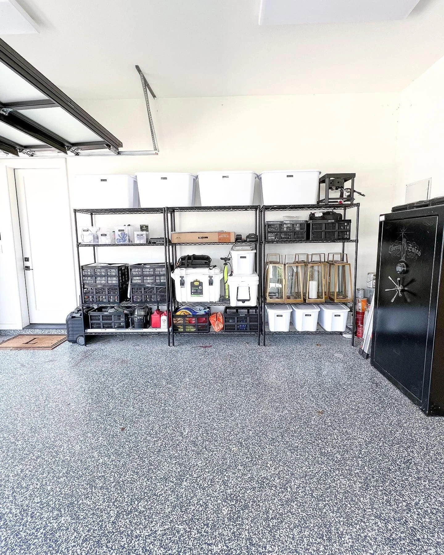 Garage storage shelving with white bins, black crates, lanterns, and various tools, next to a black safe on a speckled gray floor.