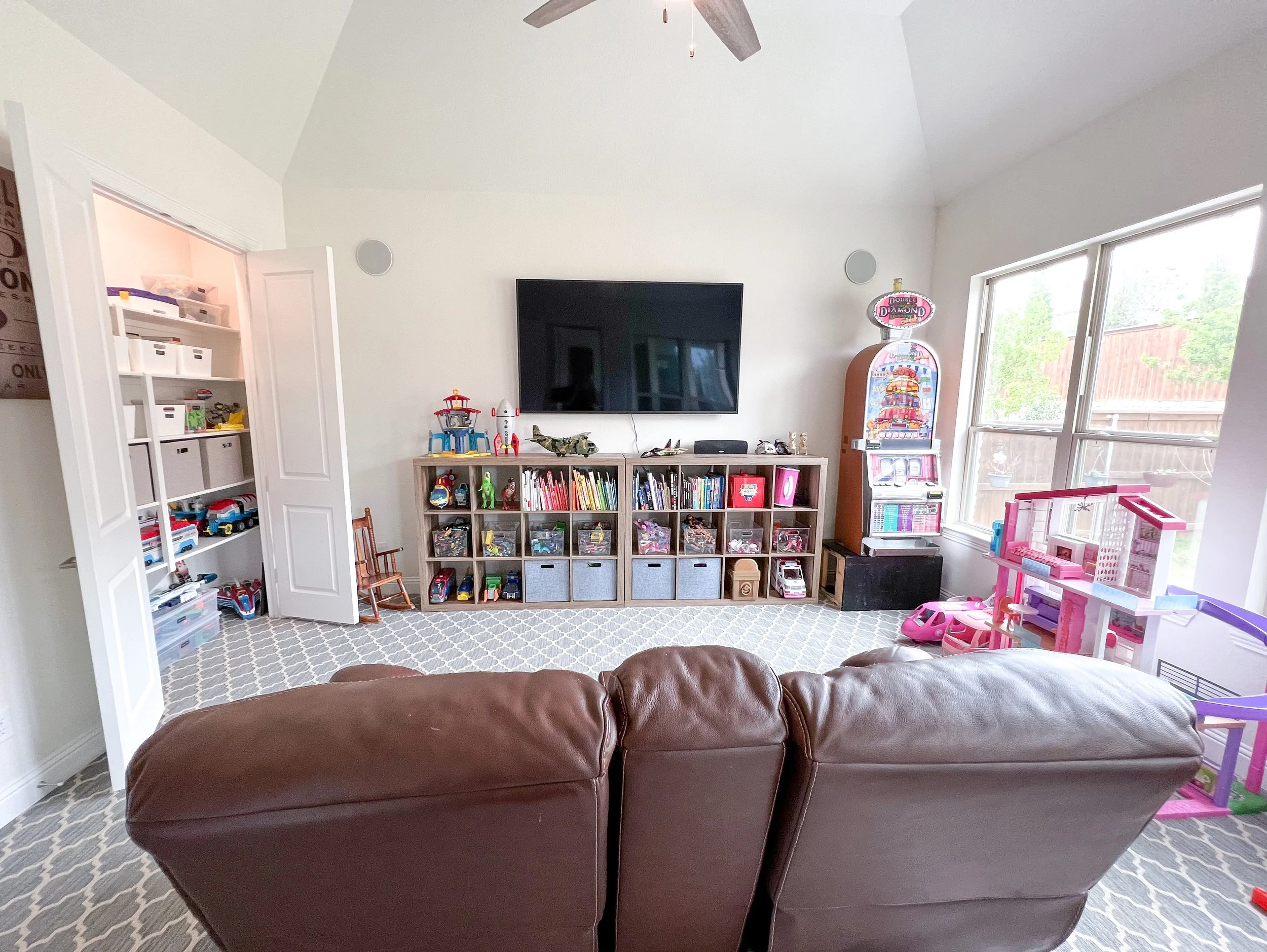 Living room with a brown leather couch, a TV mounted on the wall, a bookshelf with toys and books, a toy vending machine, a pink dollhouse, and large windows letting in natural light.
