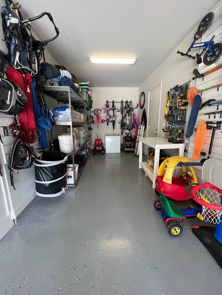 A well-organized garage with shelves holding sports gear, toys, and tools. Wall-mounted bikes and scooters, children's ride-on car and basketball hoop accessories are visible.