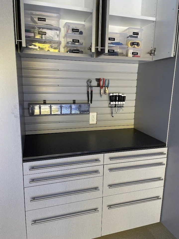 A organized workbench with lockable drawers, a black countertop, and a pegboard wall holding hand tools such as pliers, screwdrivers, and a utility knife. Upper cabinets contain labeled storage boxes for hardware and tools.