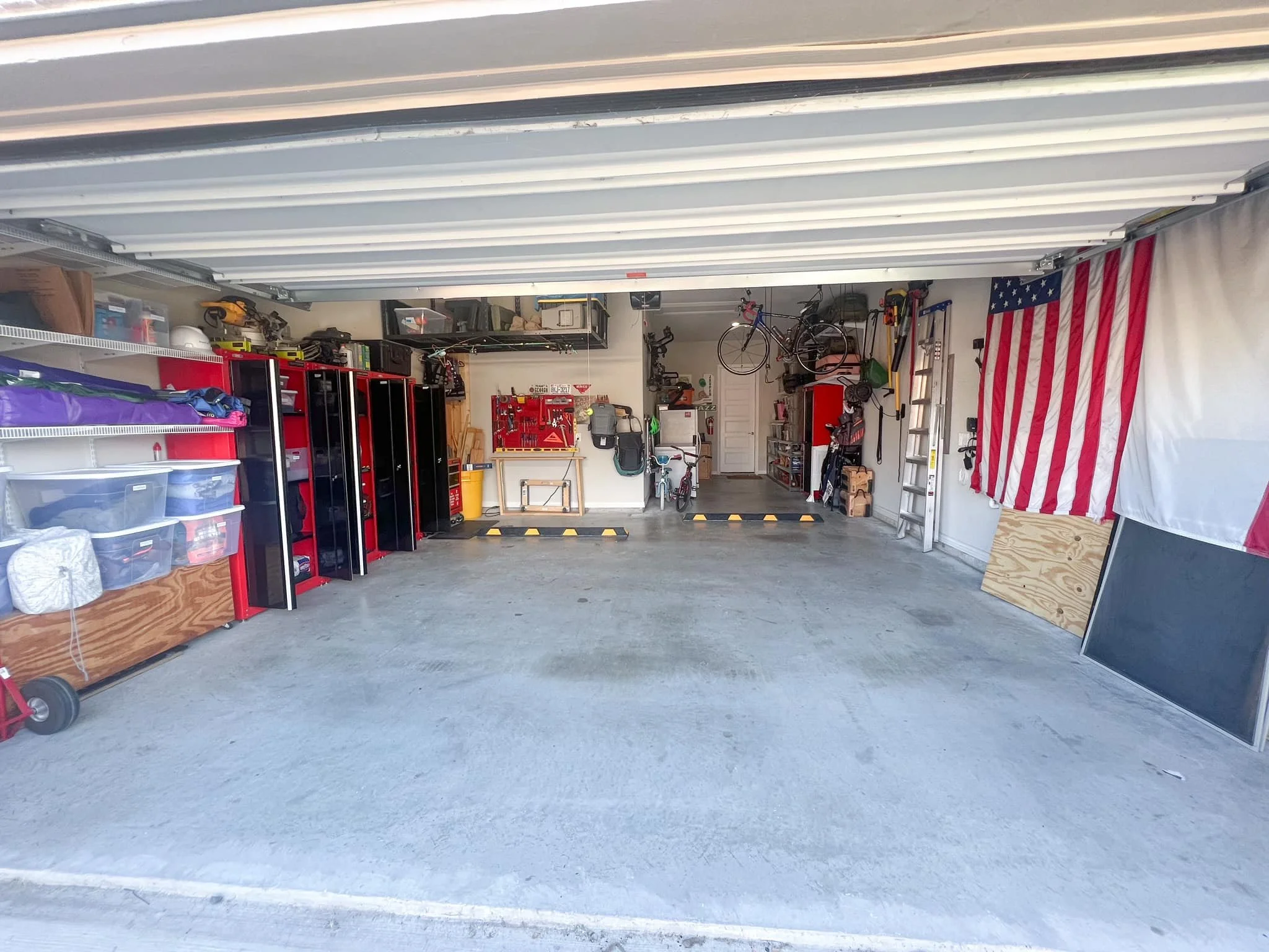 Open garage showing organized tools on red and black shelving, bicycles hanging on the wall, American flag hanging on the right side, and various stored items including a ladder, boxes, and a chalkboard leaning against the wall.