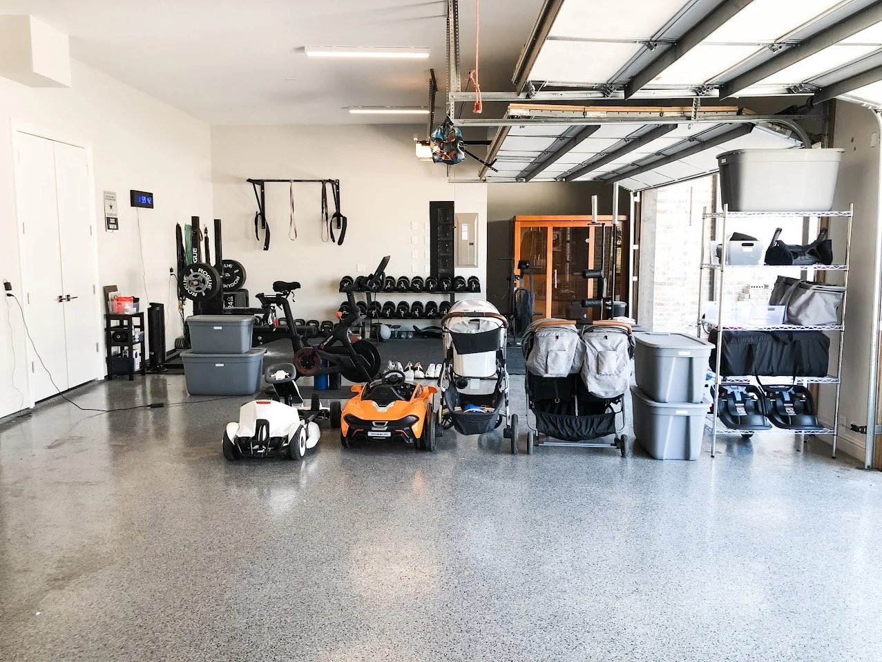 Residential garage with exercise equipment, children's ride-on toy cars, storage bins, shelving, and workout gear.