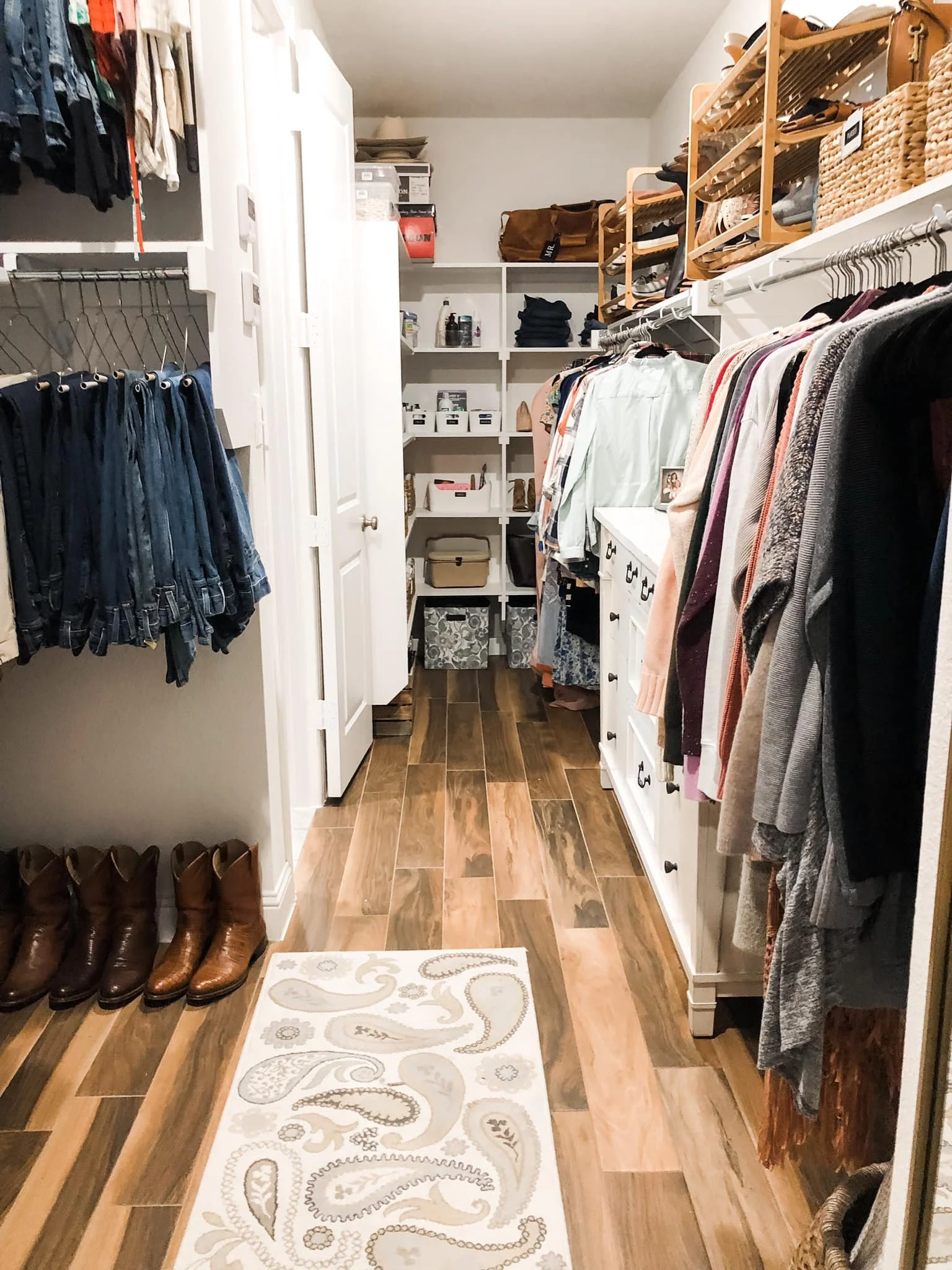 A walk-in closet with wooden flooring, white walls, and organized clothing and accessories. Left side has hanging jeans and shirts, right side has hanging sweaters and shirts, and shelves with baskets and storage boxes at the back.
