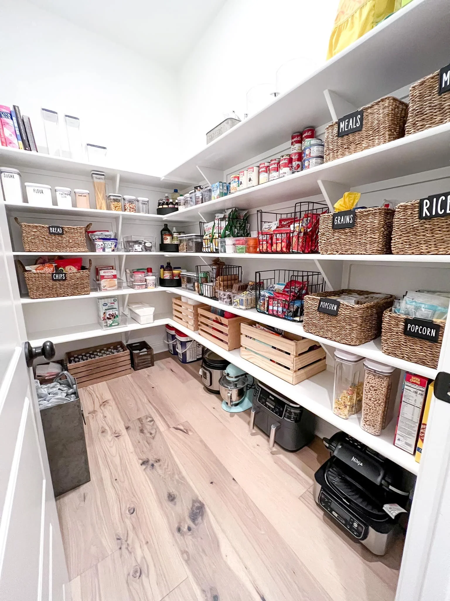 Pantry with organized white shelves labeled with black tags for bread, chips, meals, grains, rice, popcorn, with baskets, canned goods, jars, small appliances, and wooden crates on a light wood floor.