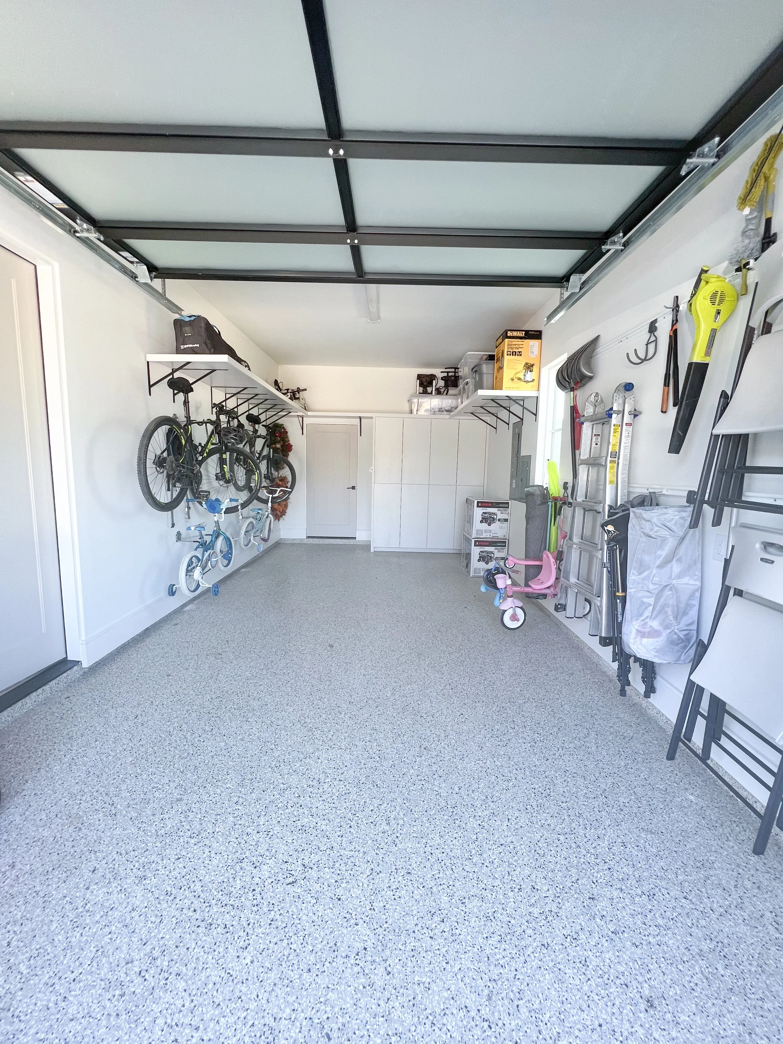 Empty garage with storage shelves, bicycles, children's tricycles, tools, and other household items organized along walls.