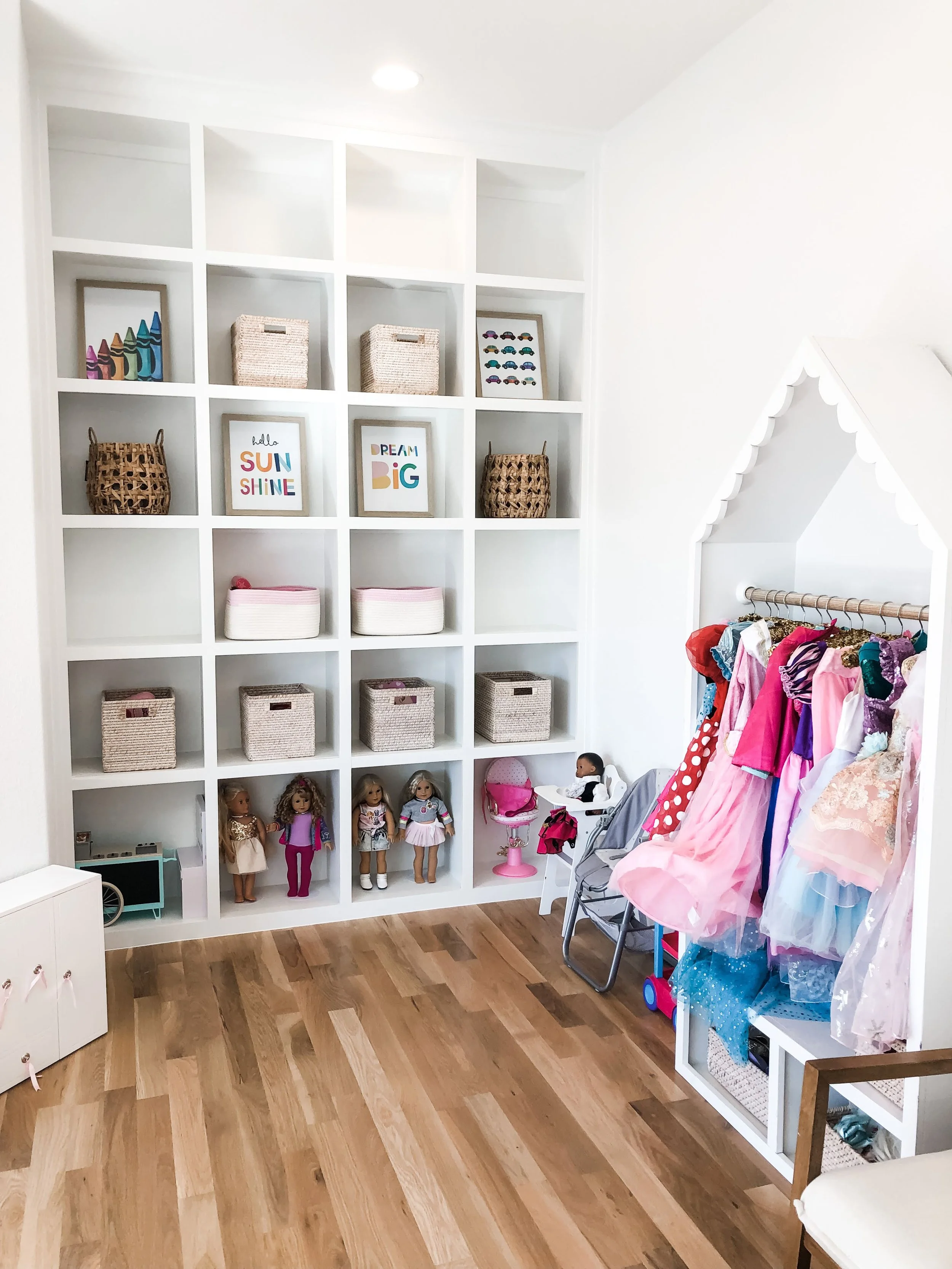 A children's room with white wall storage shelves filled with baskets, framed art, dolls, and toys. There is a small clothing rack with children's dresses on the right side, and a doll highchair, small chair, and toy chest on the floor.
